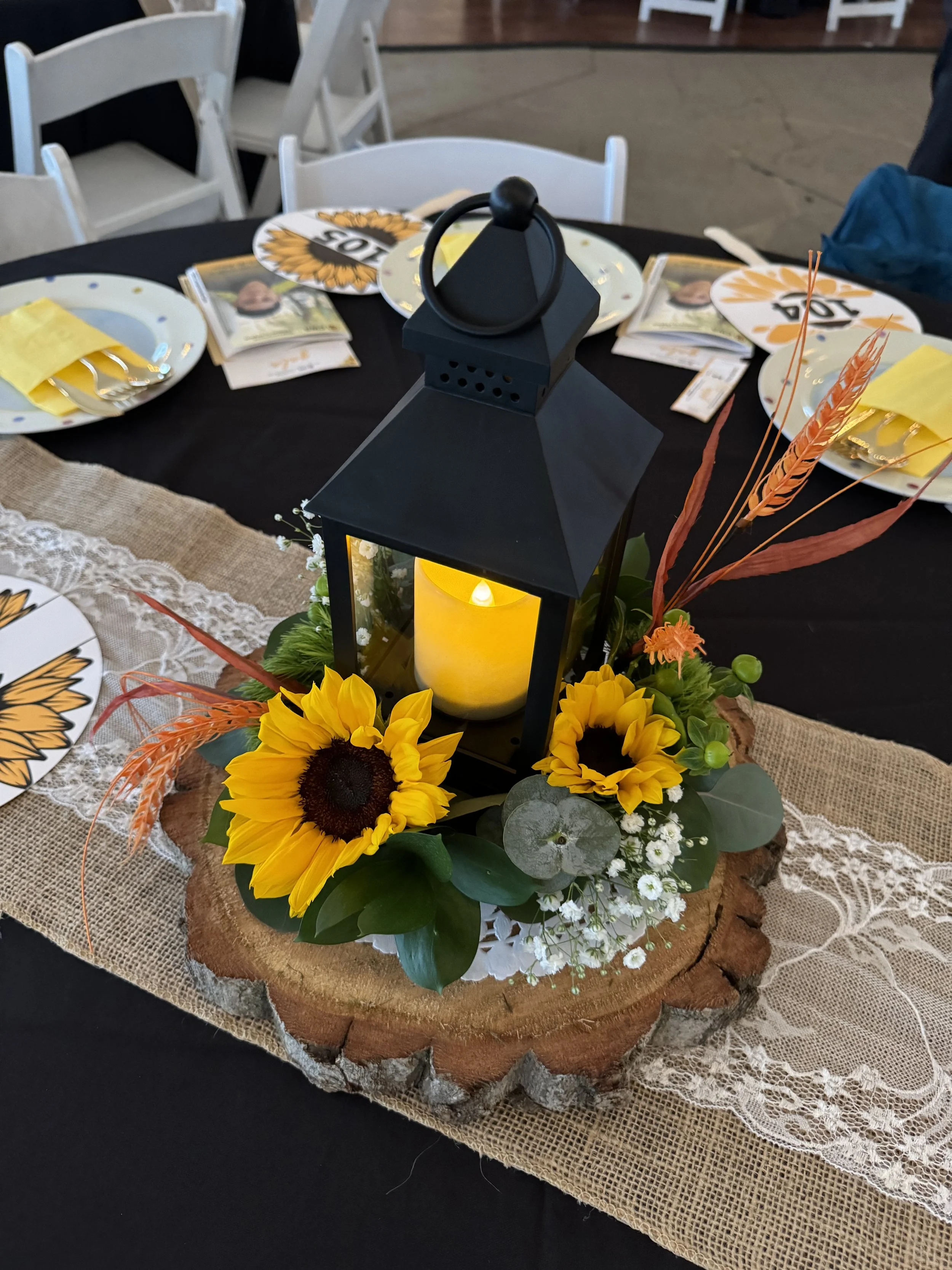 Table centerpiece with a lantern surrounded by sunflowers, greenery, and decorative wheat stalks, set on a wooden slab with lace and burlap table runner at a decorated event table.