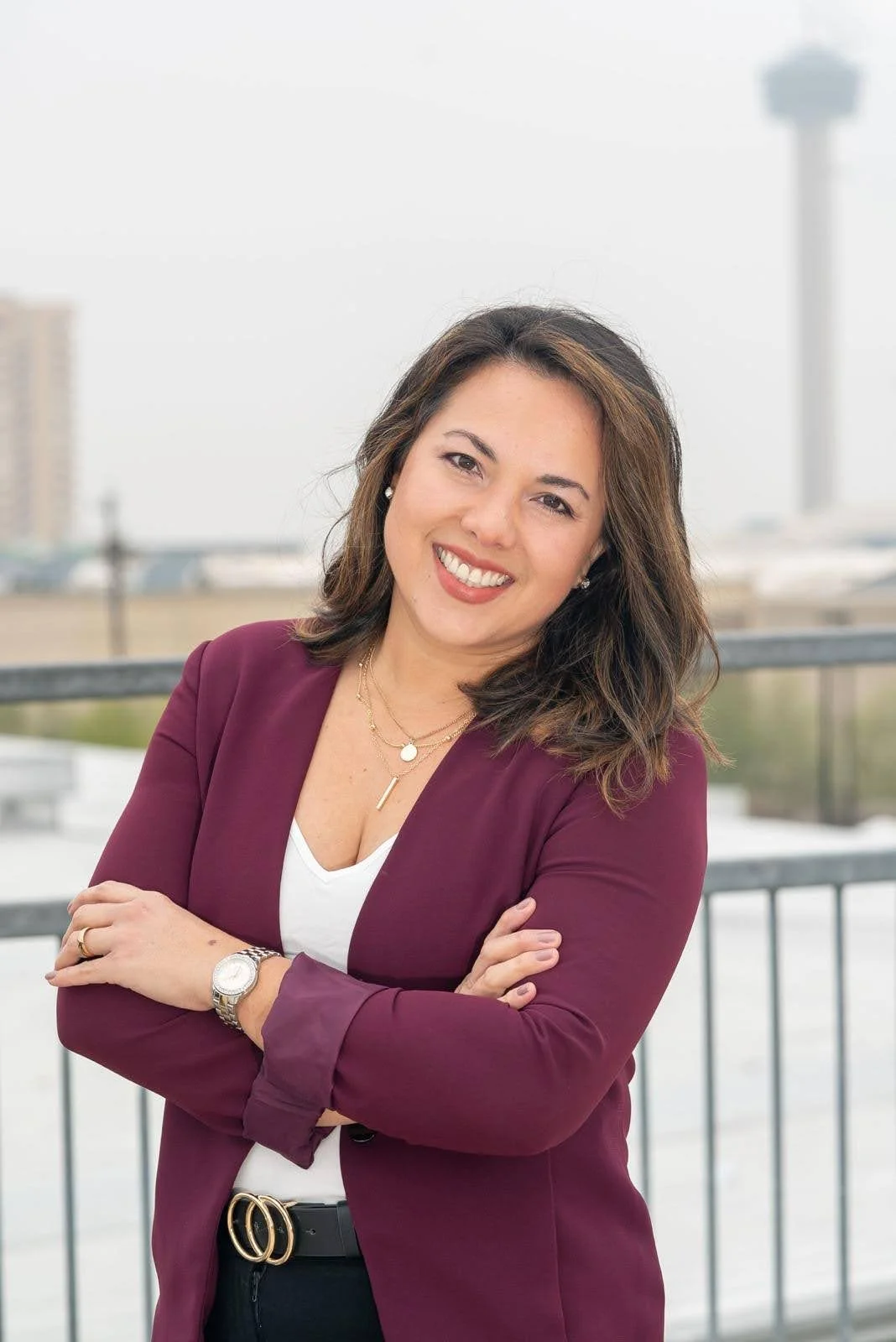 A woman with shoulder-length dark brown hair, smiling, wearing a burgundy blazer over a white top, with gold jewelry, standing outdoors with city buildings and a tower in the background.