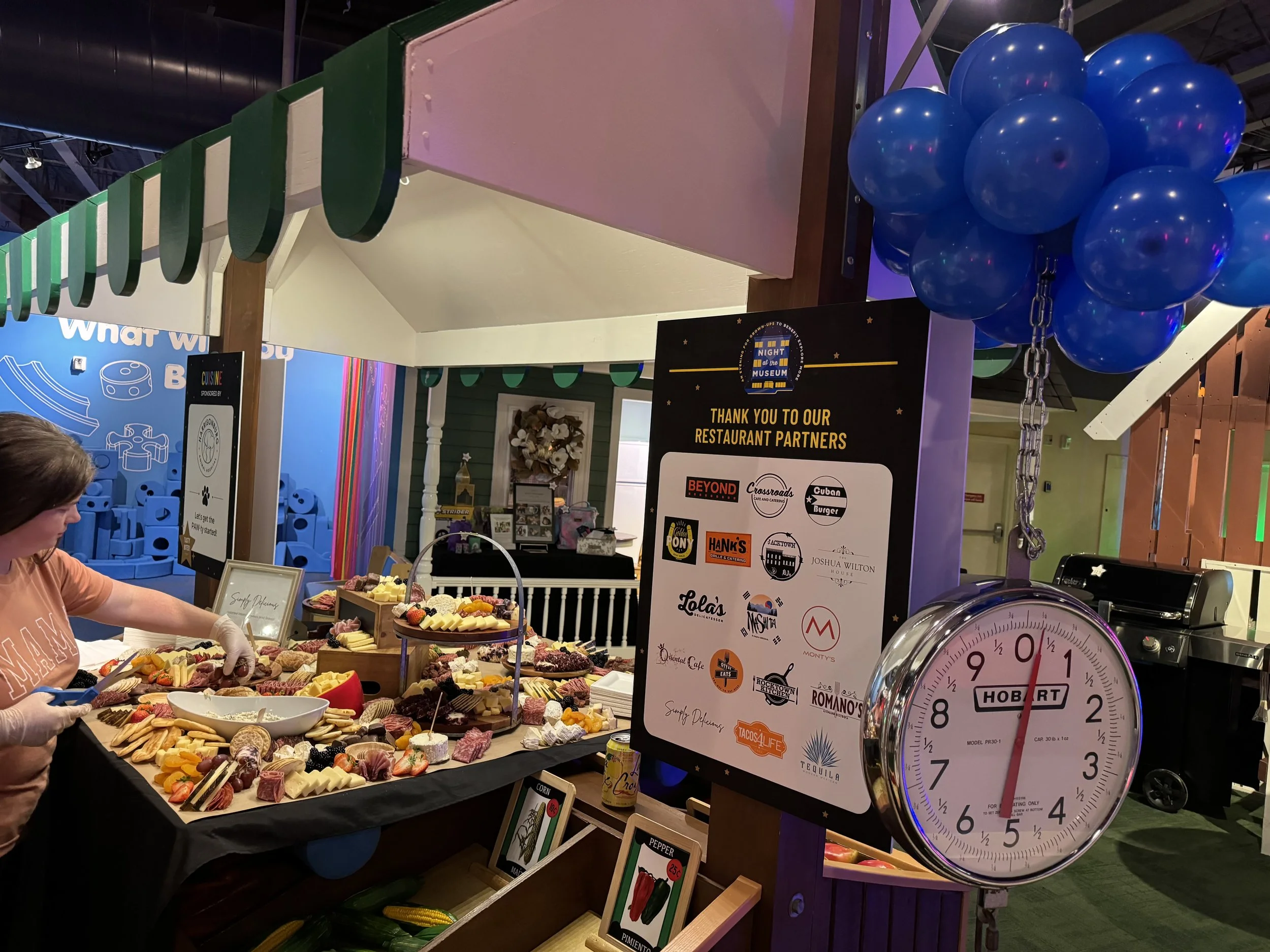 Display of assorted cheeses, meats, fruits, and snacks at a food event, with a woman preparing food, a clock showing approximately 12:05, and a sign thanking restaurant partners at a museum event.