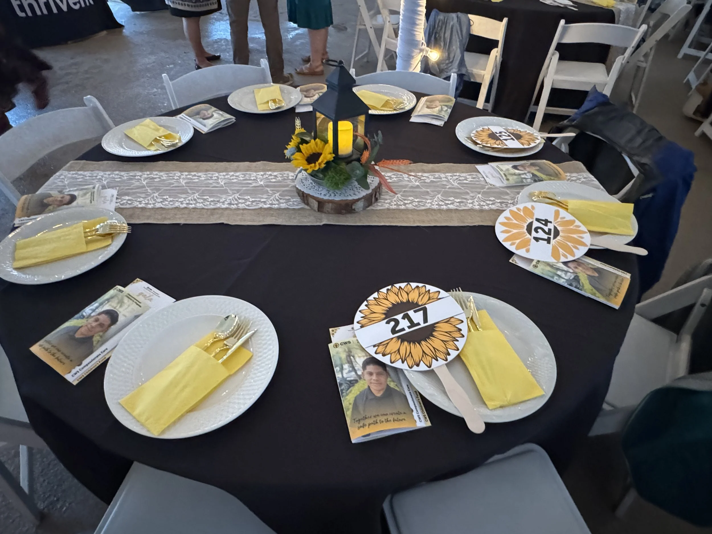 A round table set for a celebration with black tablecloth and white chairs. Place settings have white plates, yellow napkins, and gold utensils. Each setting has a photo card and a paper plate with a sunflower design and a number, including 217, 124, and 123. The table centerpiece is a lantern with a candle inside, surrounded by sunflowers, green leaves, and decorative orange feathers.