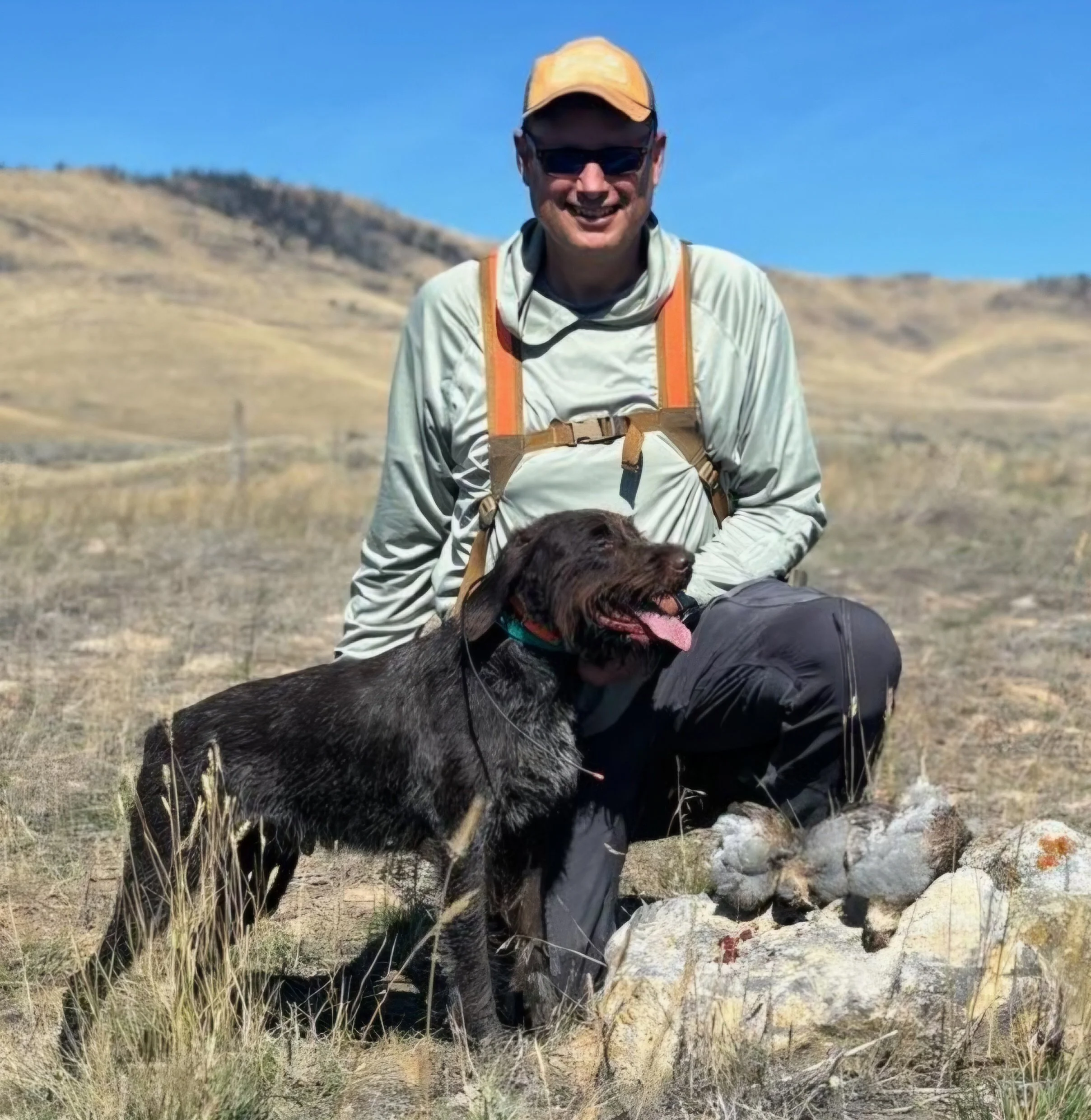 A man in outdoor gear smiling at the camera, kneeling with a chocolate Labrador retriever in an open grassy field with rolling hills in the background.