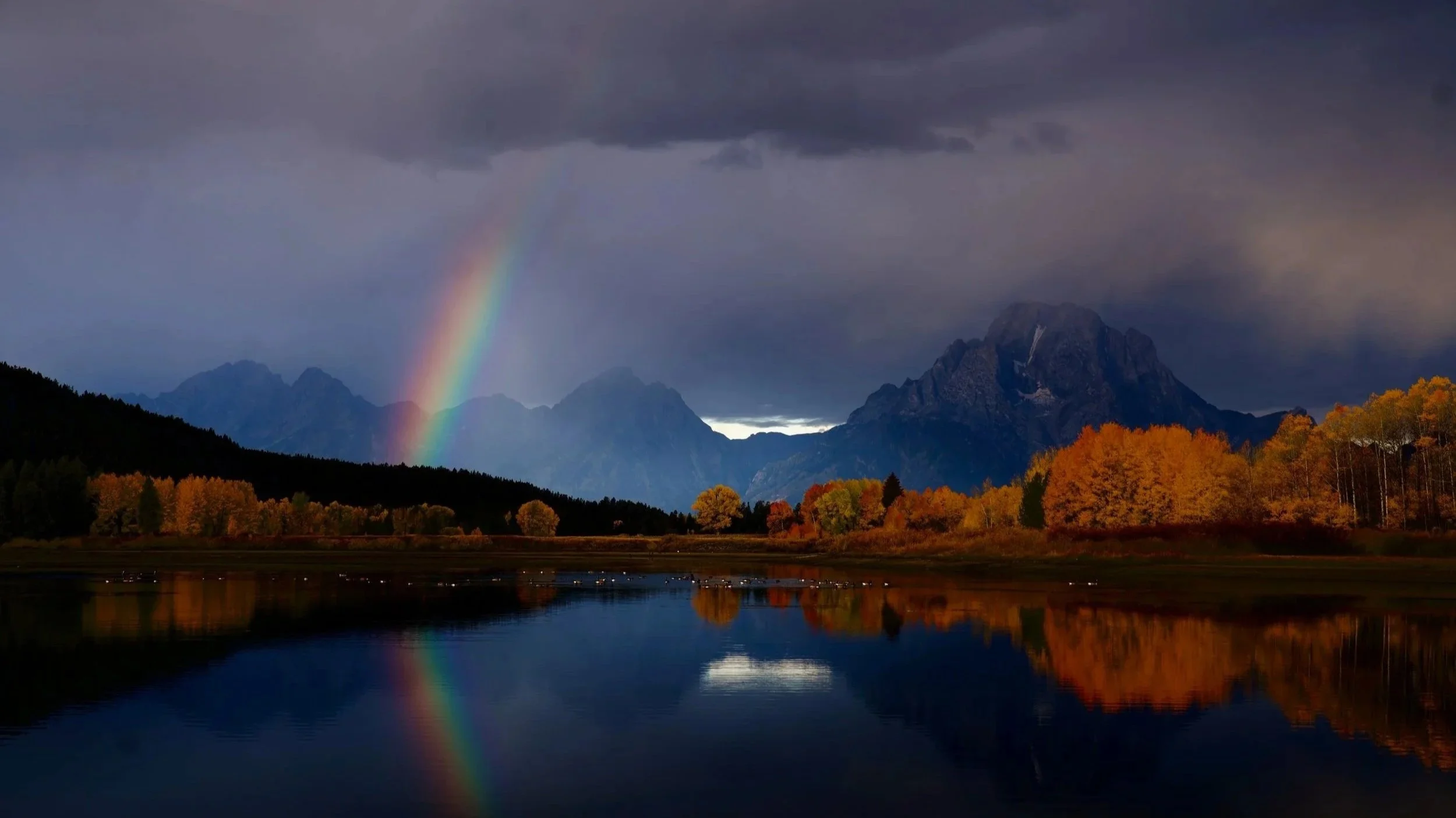 A landscape scene with a rainbow in the cloudy sky, mountains in the background, and colorful trees reflected in a calm lake.