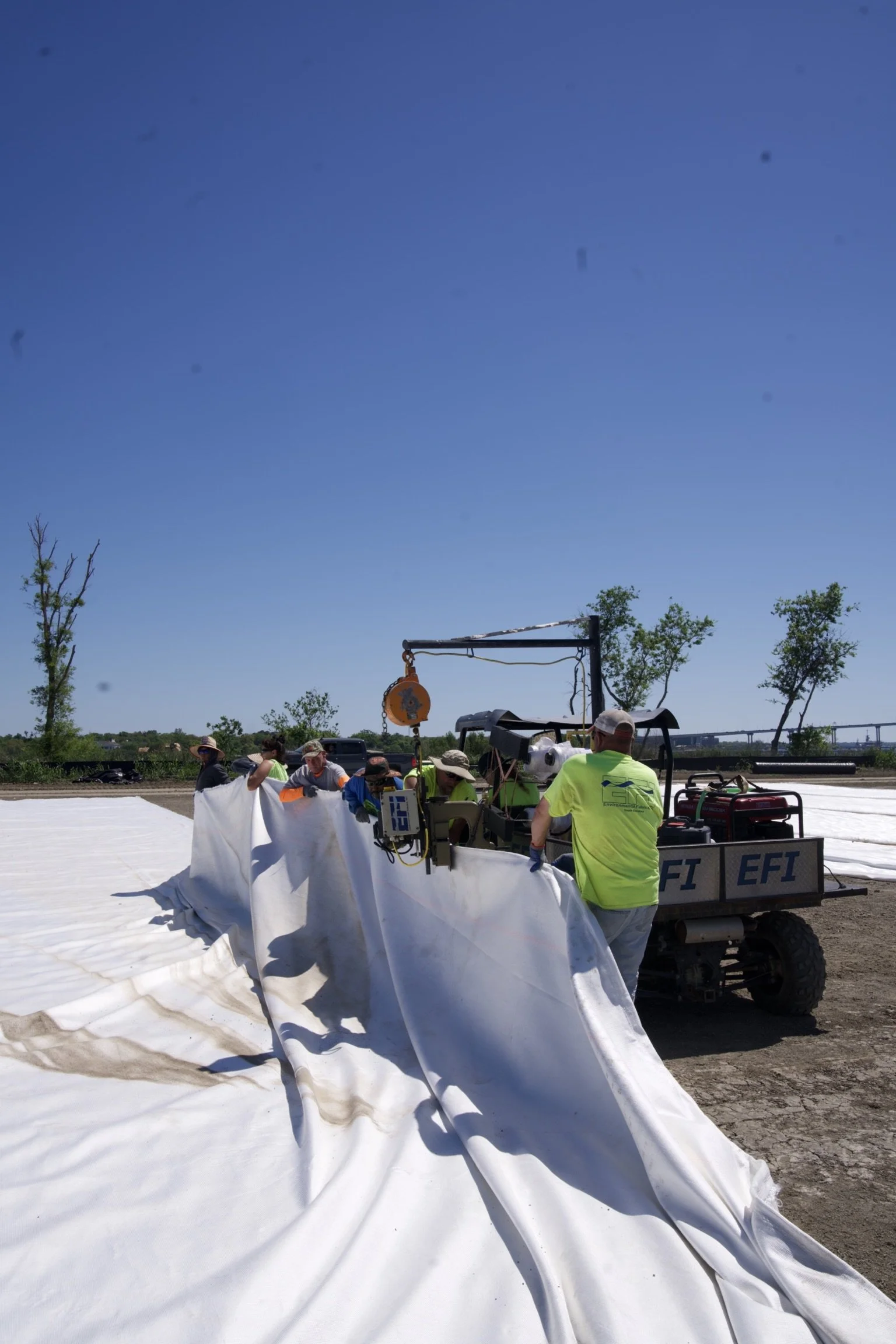 A group of workers laying out large white fabric on the ground outdoors under a clear blue sky, with some trees in the background.