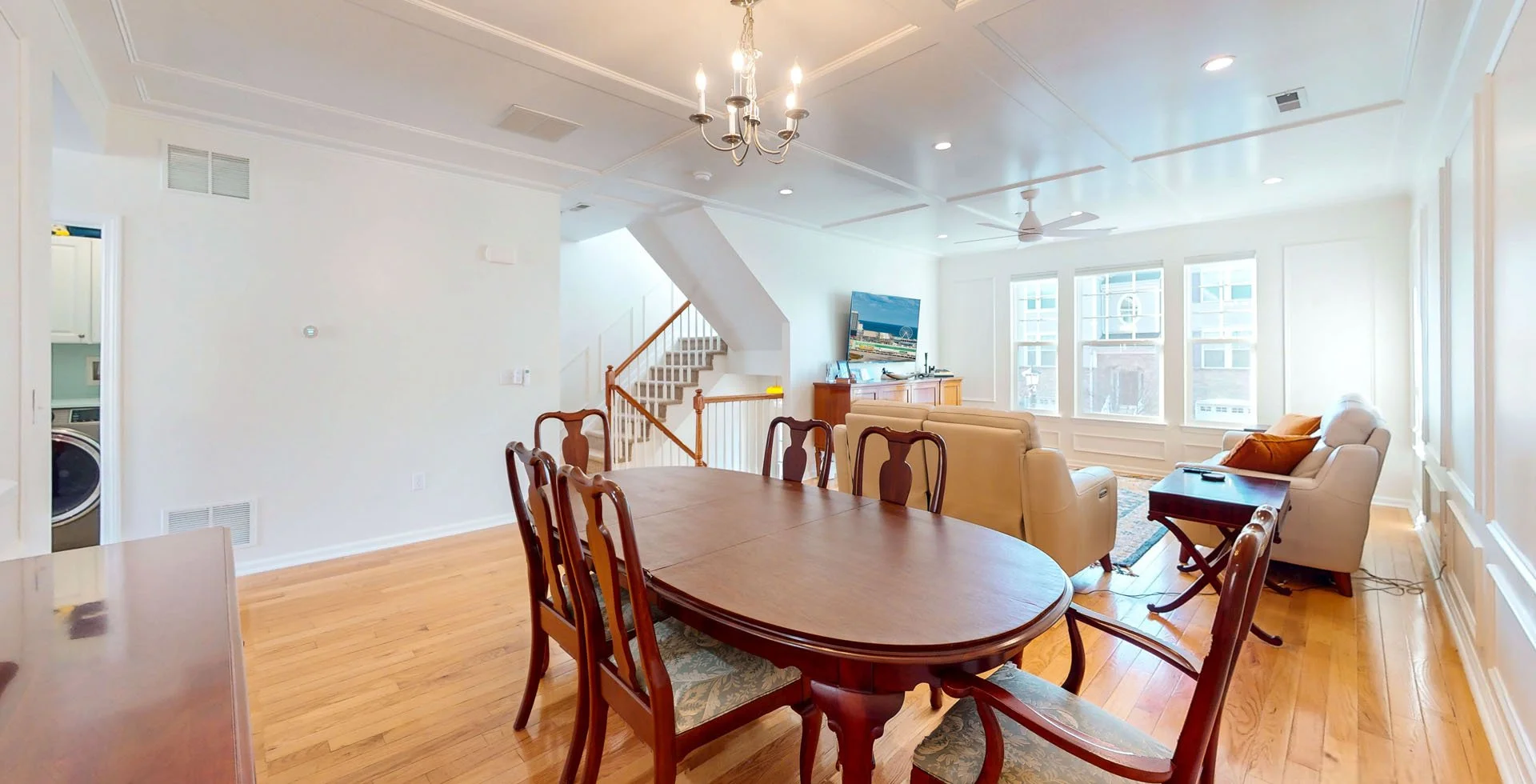 Bright living room with large windows, hardwood floors, a wooden dining table with chairs, a beige sofa with cushions, a TV on a stand, and staircase with wooden railing.