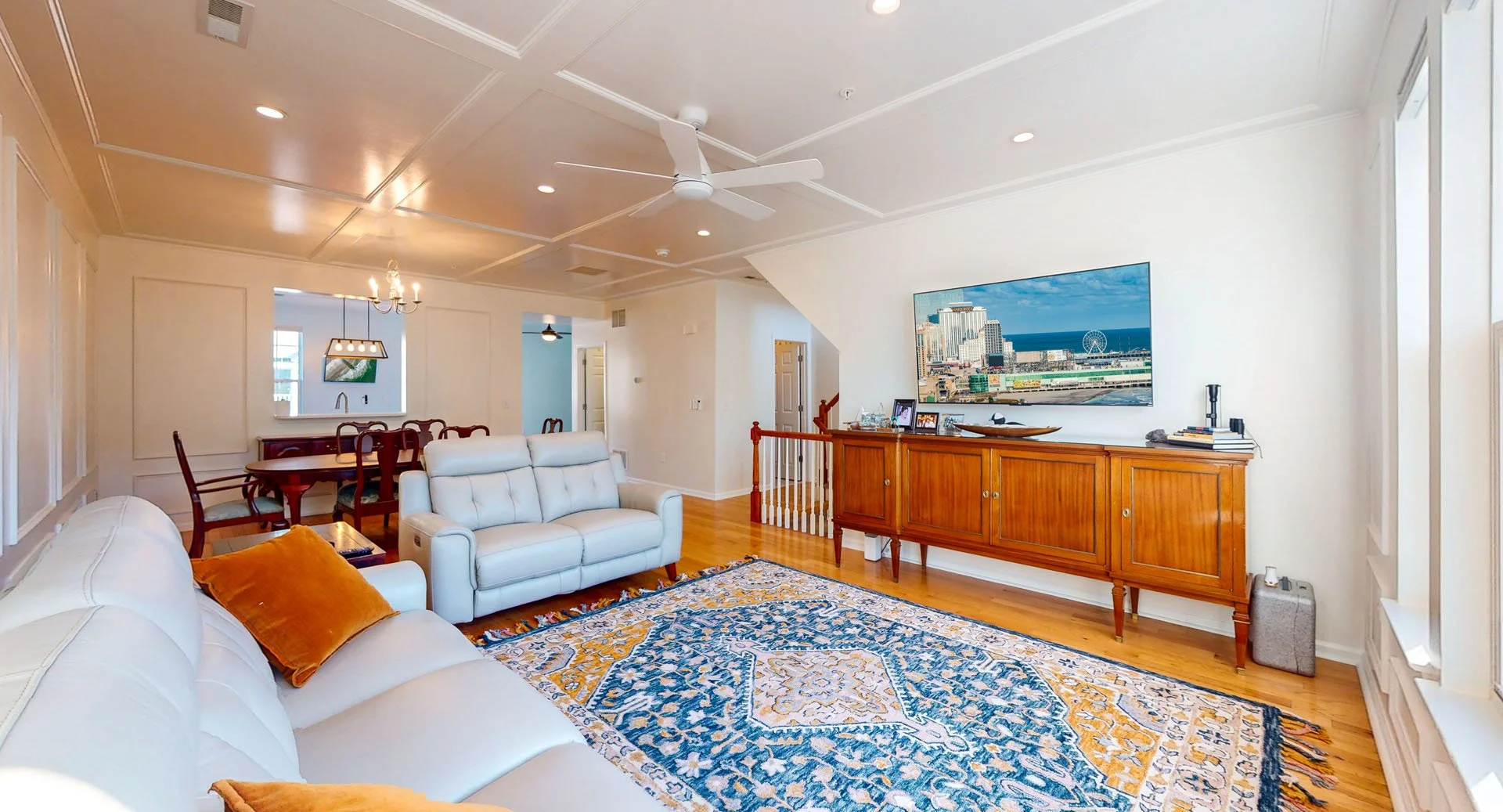 Living room with hardwood floors, white walls, a large blue and yellow patterned rug, white sofas with orange cushions, a wooden console with a TV, and a dining area with a wooden table and chairs.