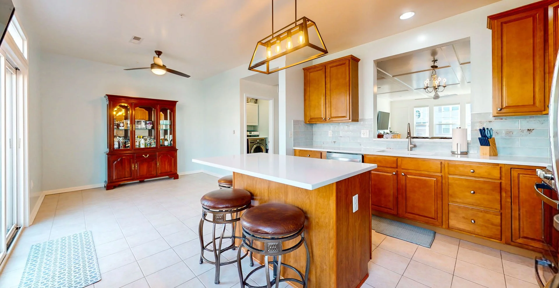 Kitchen with wooden cabinets, white countertops, and a view into a laundry room with a washing machine. There is an island with three bar stools, a hanging light fixture, and a sliding glass door leading outside. A red china cabinet is against the wa