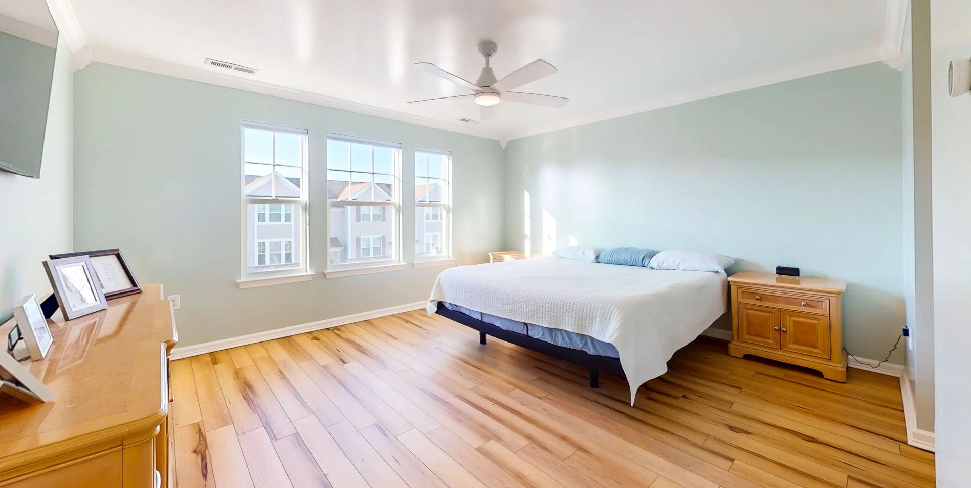A bedroom with a bed, wooden nightstands, a dresser with picture frames, windows letting in natural light, and a ceiling fan.