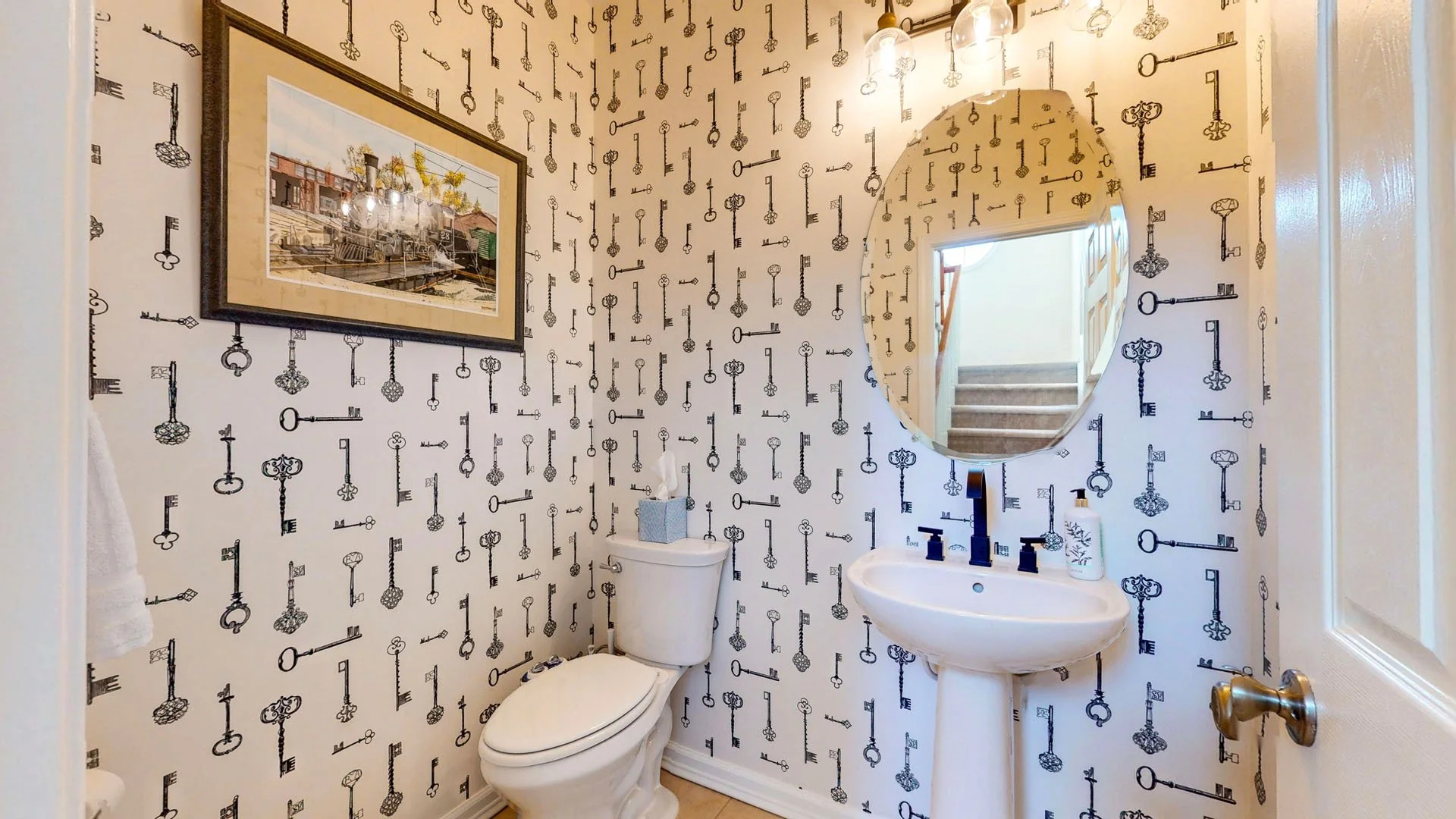 Small bathroom with wallpaper featuring various black antique keys, white toilet, pedestal sink with black faucet, oval mirror, framed picture on the wall, tissue box on the toilet, and a bottle of lotion on the sink.