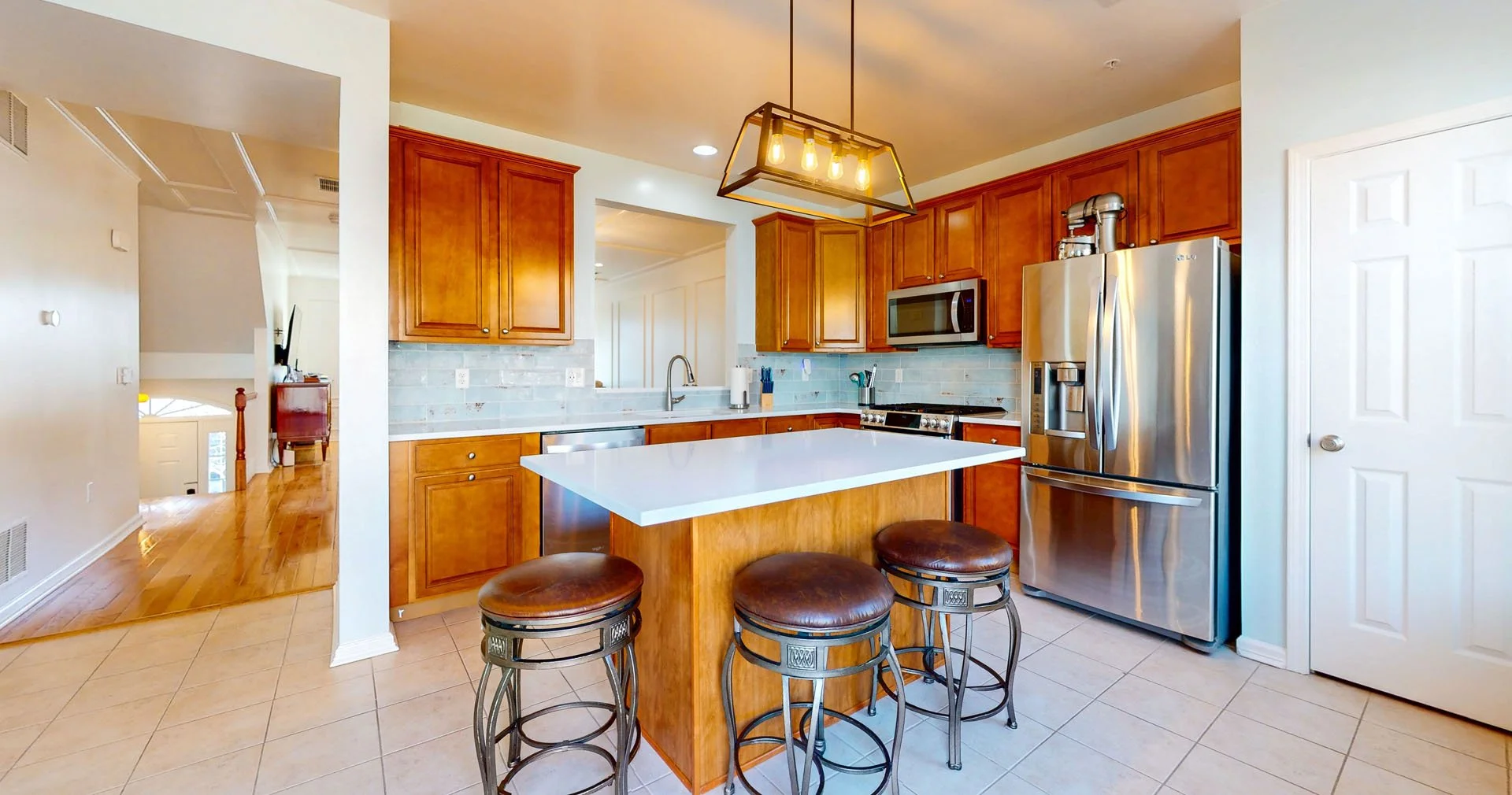 Kitchen with wooden cabinets, stainless steel refrigerator, microwave, and island with three bar stools, pendant light, and tiled floor.