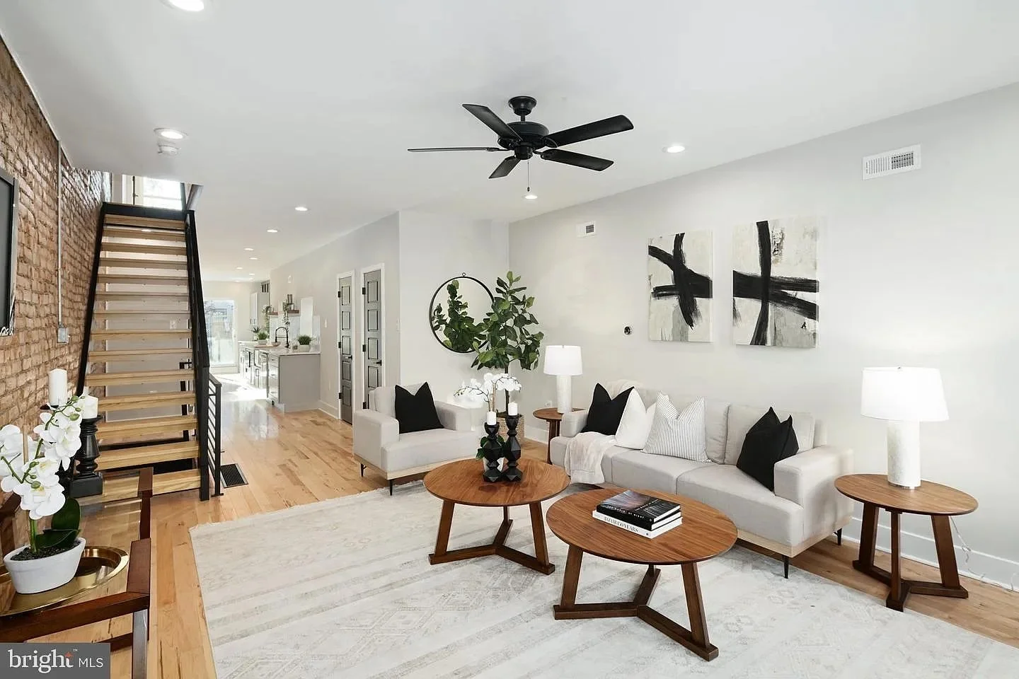 Bright living room with white sofa, black and white pillows, wooden coffee tables, lamp, and artwork, with staircase on the left and hardwood floors.
