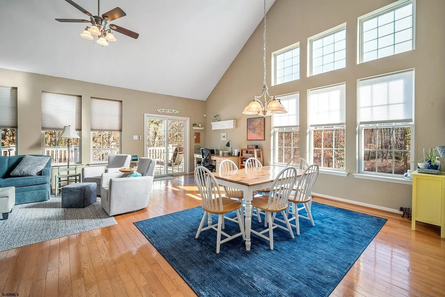 Bright living room with hardwood floors, beige walls, a blue area rug, a round wooden dining table with six white chairs, large windows, a ceiling fan, and a desk area with a chair and shelves.
