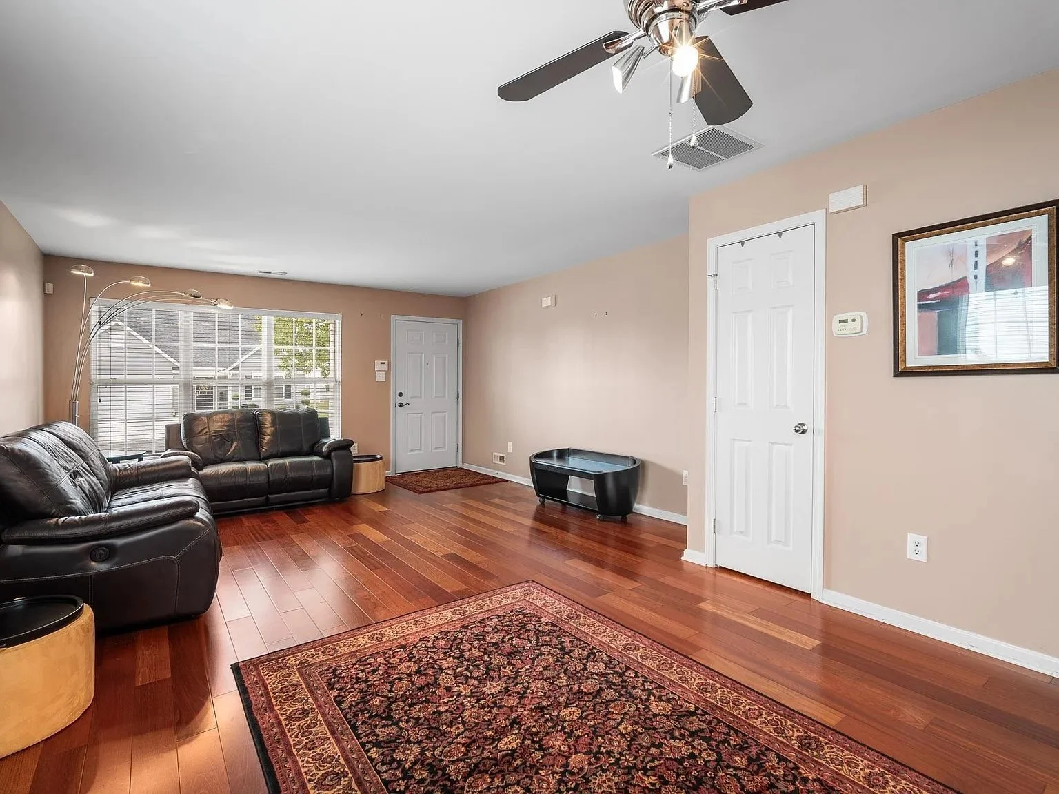 Living room with hardwood floors, beige walls, a dark leather sofa set, a large window with blinds, a colorful area rug, a ceiling fan with lights, a framed picture on the wall, and a small black storage bench.