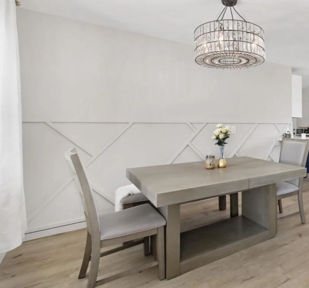 A dining room with a rectangular wooden table, two chairs, a chandelier, a small vase with white flowers, and a wall with a geometric design.