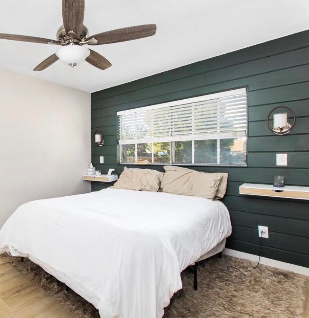 A bedroom with a bed, beige pillows, dark green shiplap accent wall, large window with white blinds, floating shelves with decor, ceiling fan, and beige rug.
