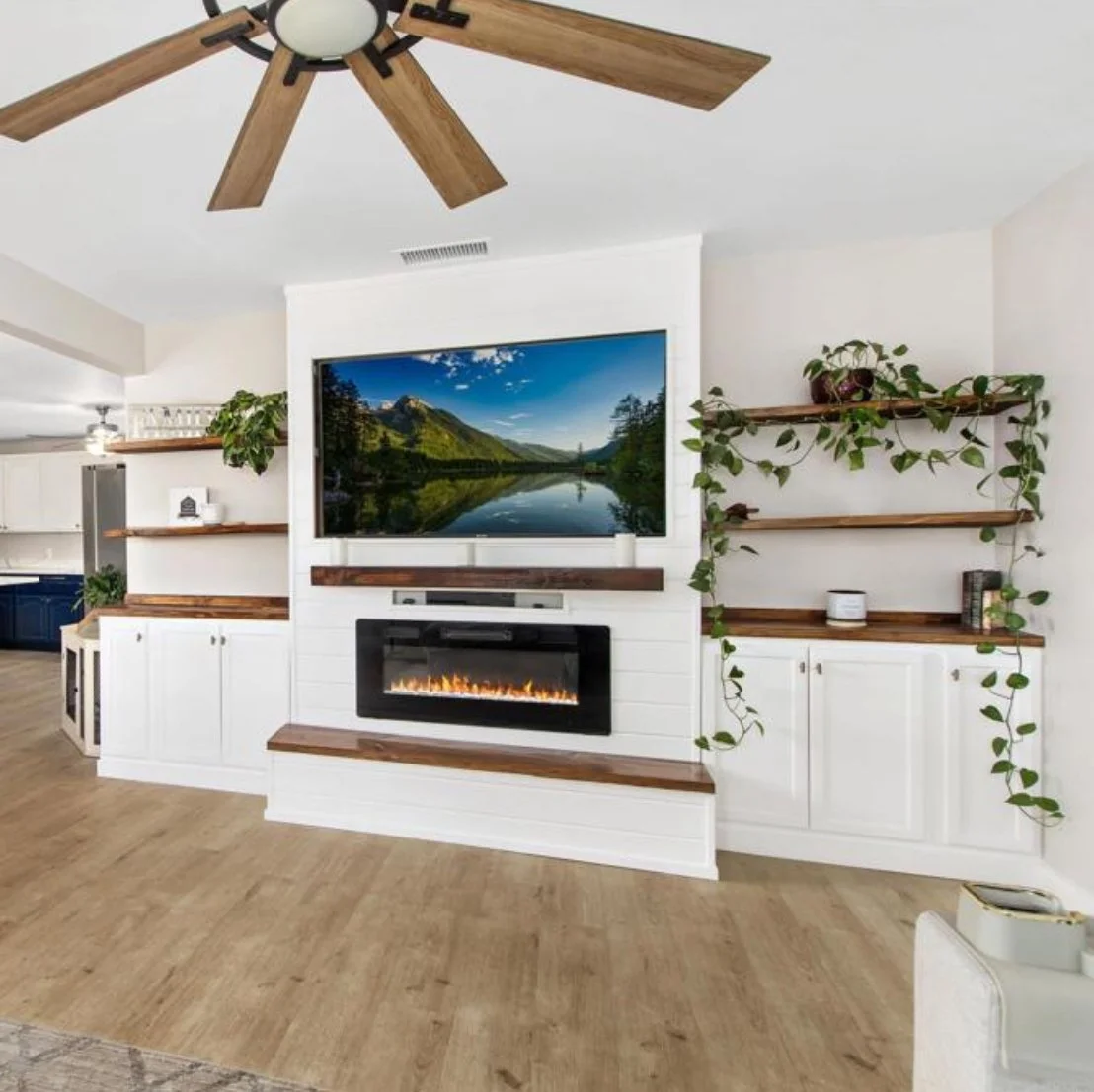 Living room with a modern electric fireplace below a large flat-screen TV displaying nature scenery, surrounded by white shelves with plants and decorative items, wooden accents, a ceiling fan, and light wood flooring.