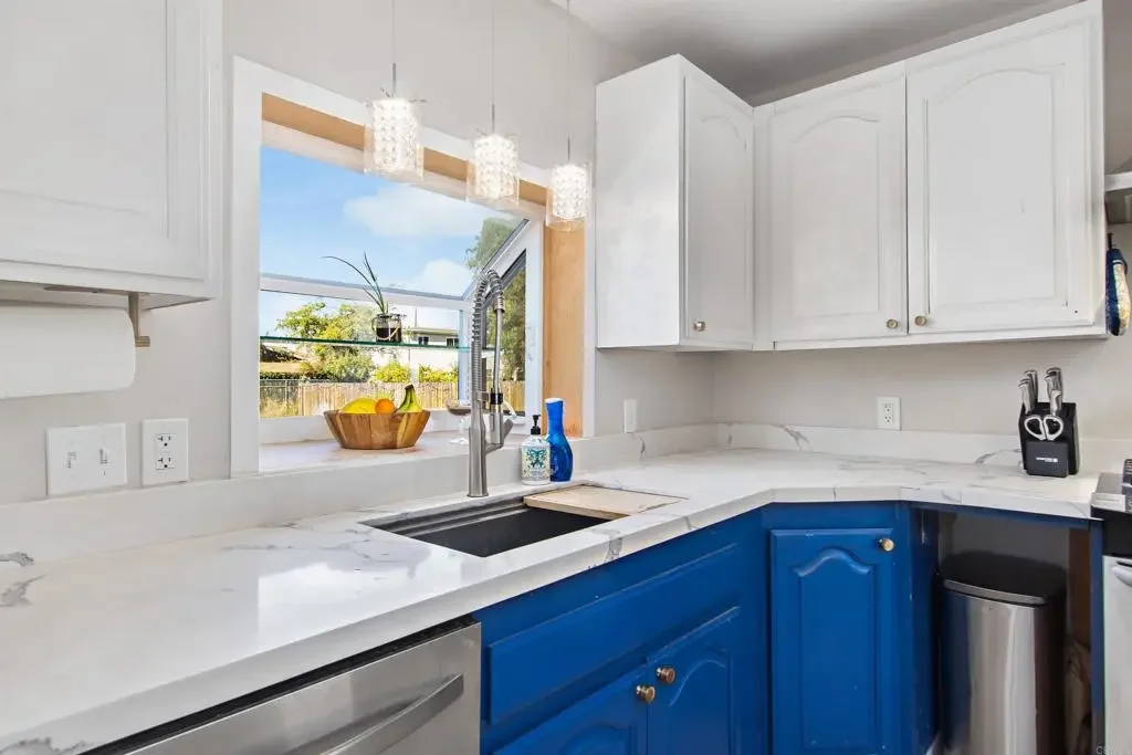 Modern kitchen with white upper cabinets, blue lower cabinets, marble countertop, and a window with a view of a backyard garden.