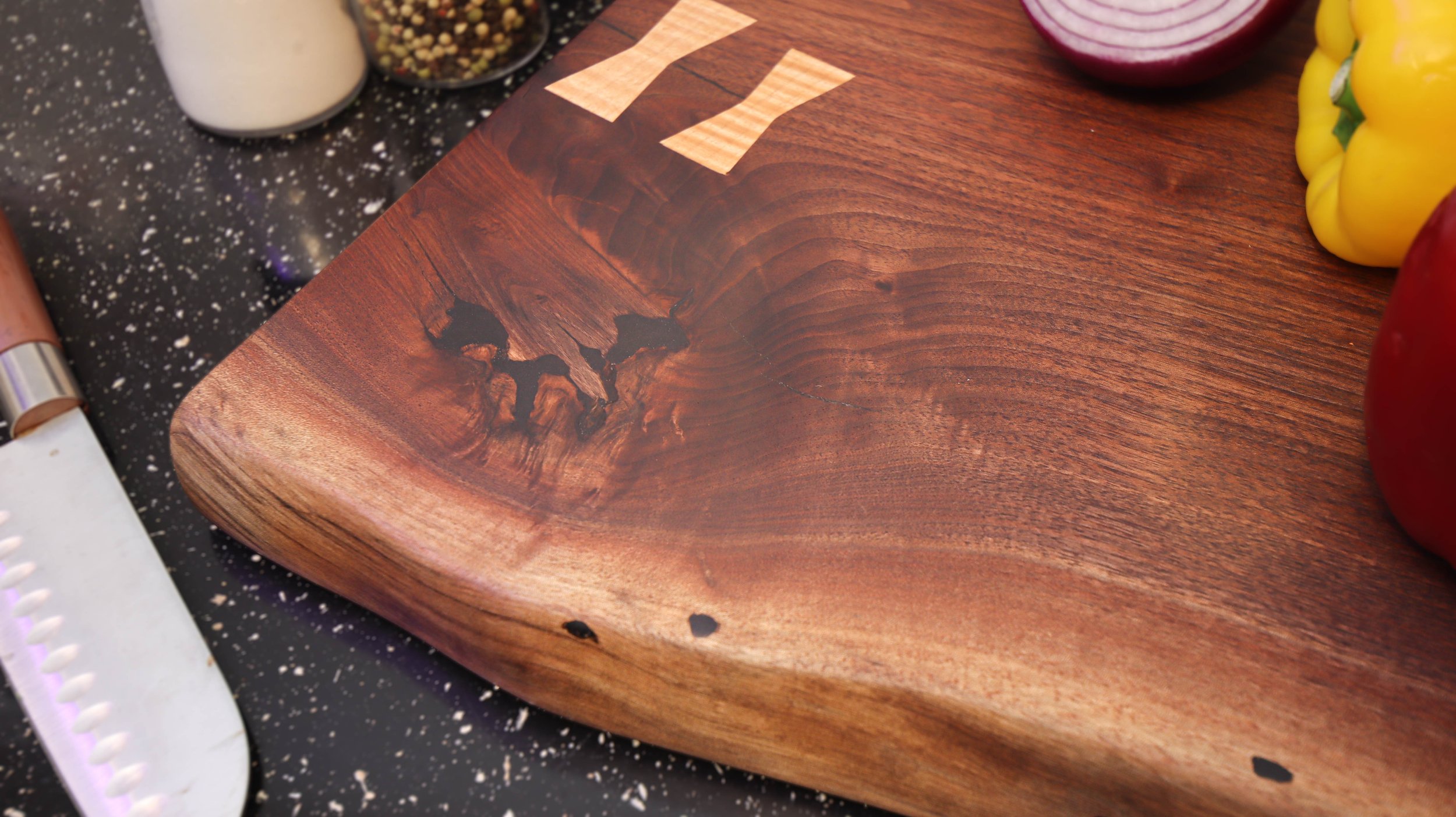 Close-up of a wooden cutting board with a decorative knot in the wood grain, placed on a black speckled countertop with peppers, onions, and spices around it.