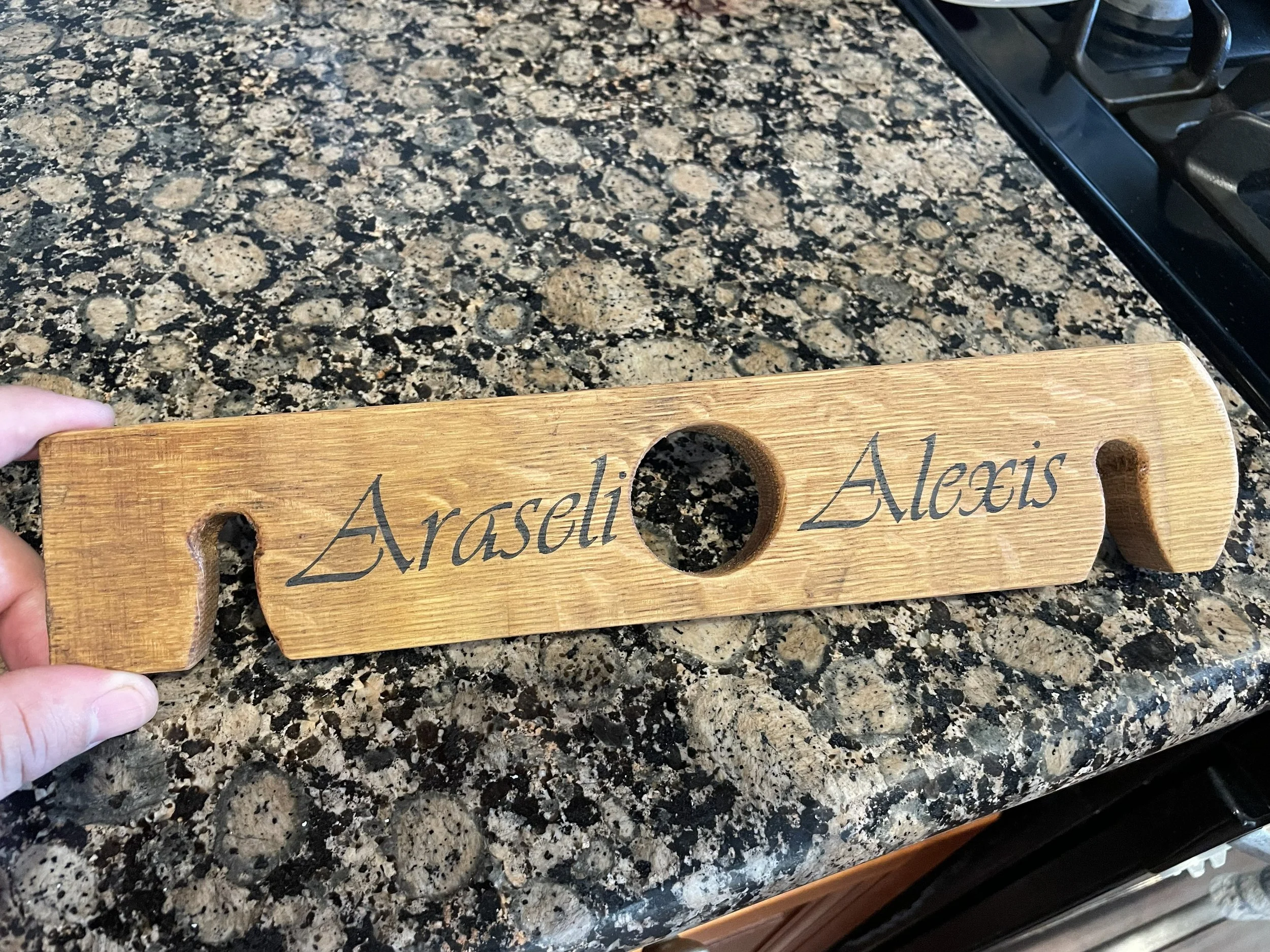 A barrel stave wine caddy personalized with the names "Araseli Alexis" engraved on it, held by someone's hand, resting on a granite kitchen countertop.