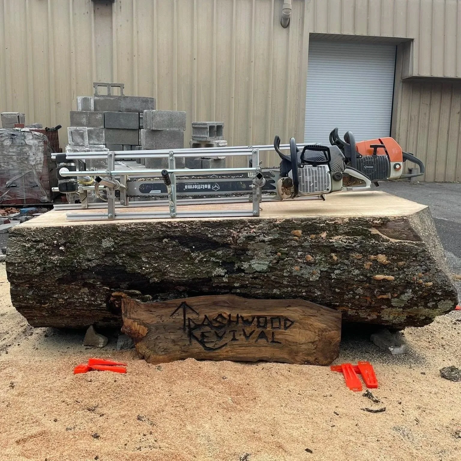 A large red oak log with a wooden sign that says "Trashwood Revival" on it. On top of the stump, there are chainsaws and a sawmill, with a background of a metal building, cement blocks, and a garage door.