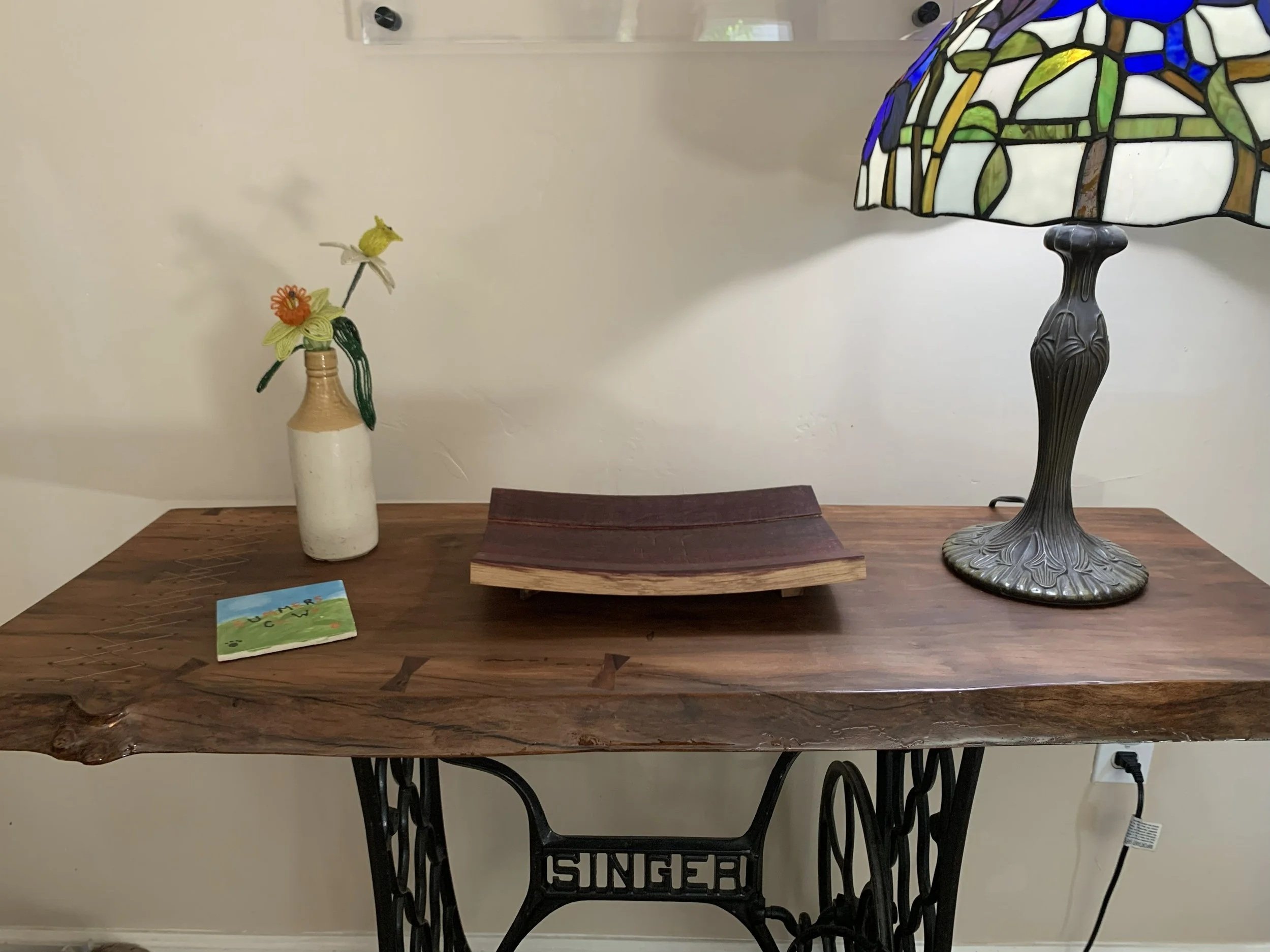 A rustic wooden table with decorative items including a ceramic vase with artificial flowers, a vintage-style lamp with a stained glass shade, and a wooden tray, against a plain wall in a room.