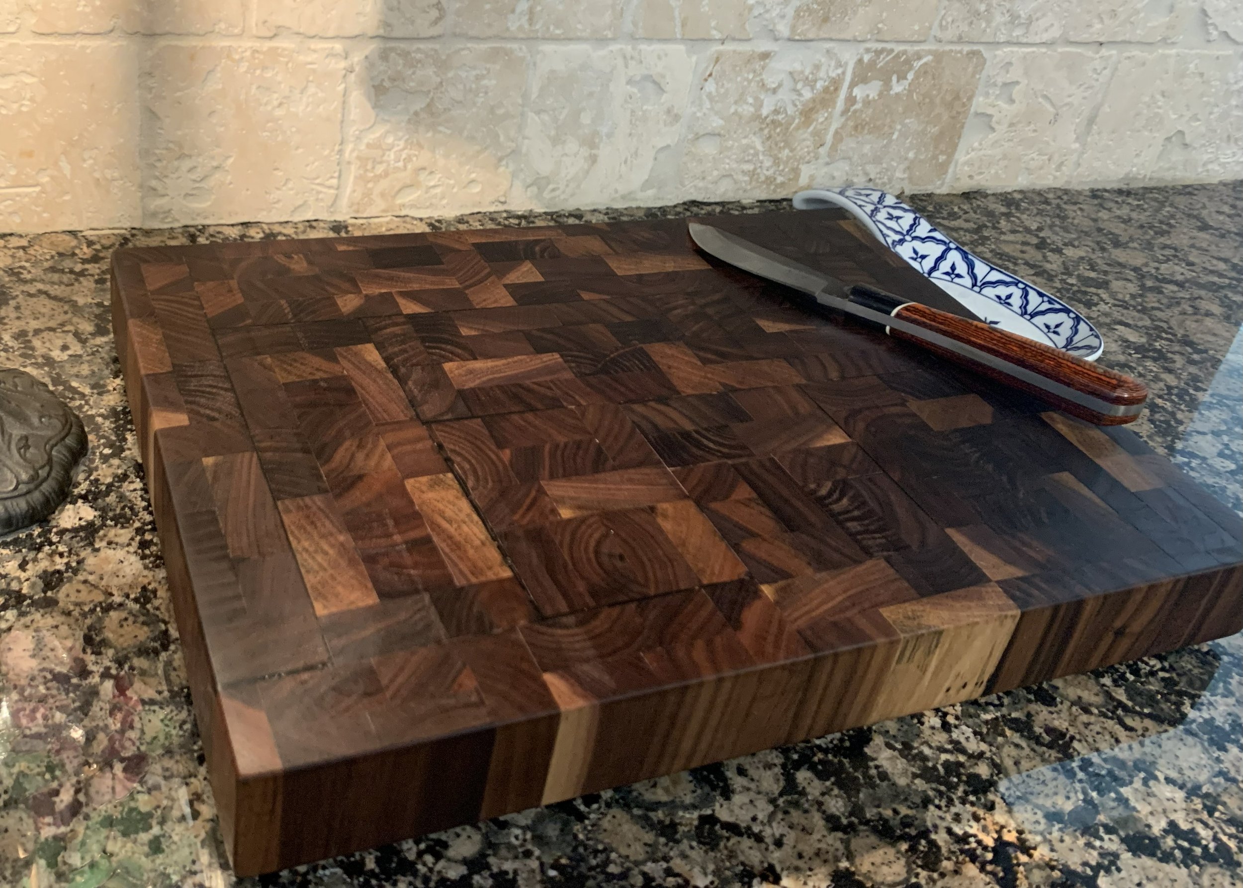 A wooden cutting board with a pattern of various kinds of wood inlay, a knife with a wooden handle, and a decorative cloth on a granite kitchen countertop.