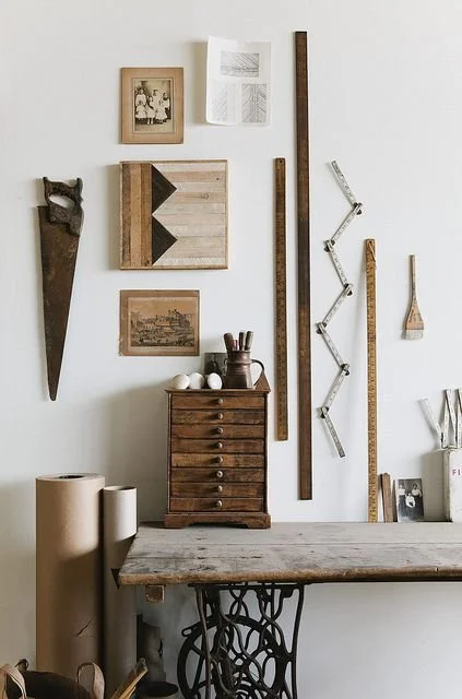 Interior view of a room with a rustic wooden table, a small wooden drawer unit, various vintage tools and rulers on the wall, framed photographs and artwork, and cardboard tubes on the floor.