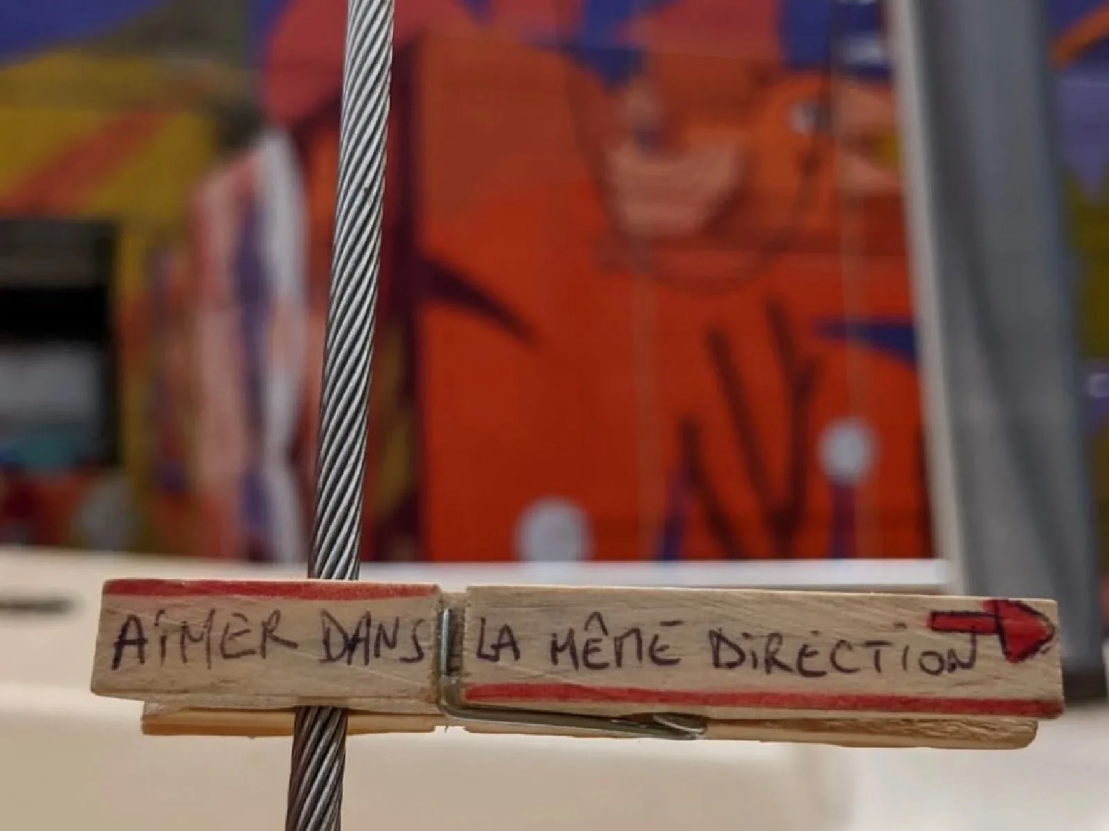 A small wooden stick with a handwritten message in French that says 'Aimér dans la même direction' with an arrow pointing to the right, attached to a metallic wire, with a blurred colorful fabric background. The photo is taken in Bordeaux, France.