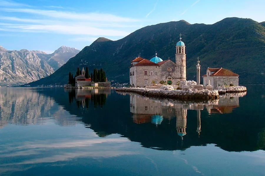 A small church on an island in a calm lake, surrounded by mountains, with clear blue skies, located in Montenegro