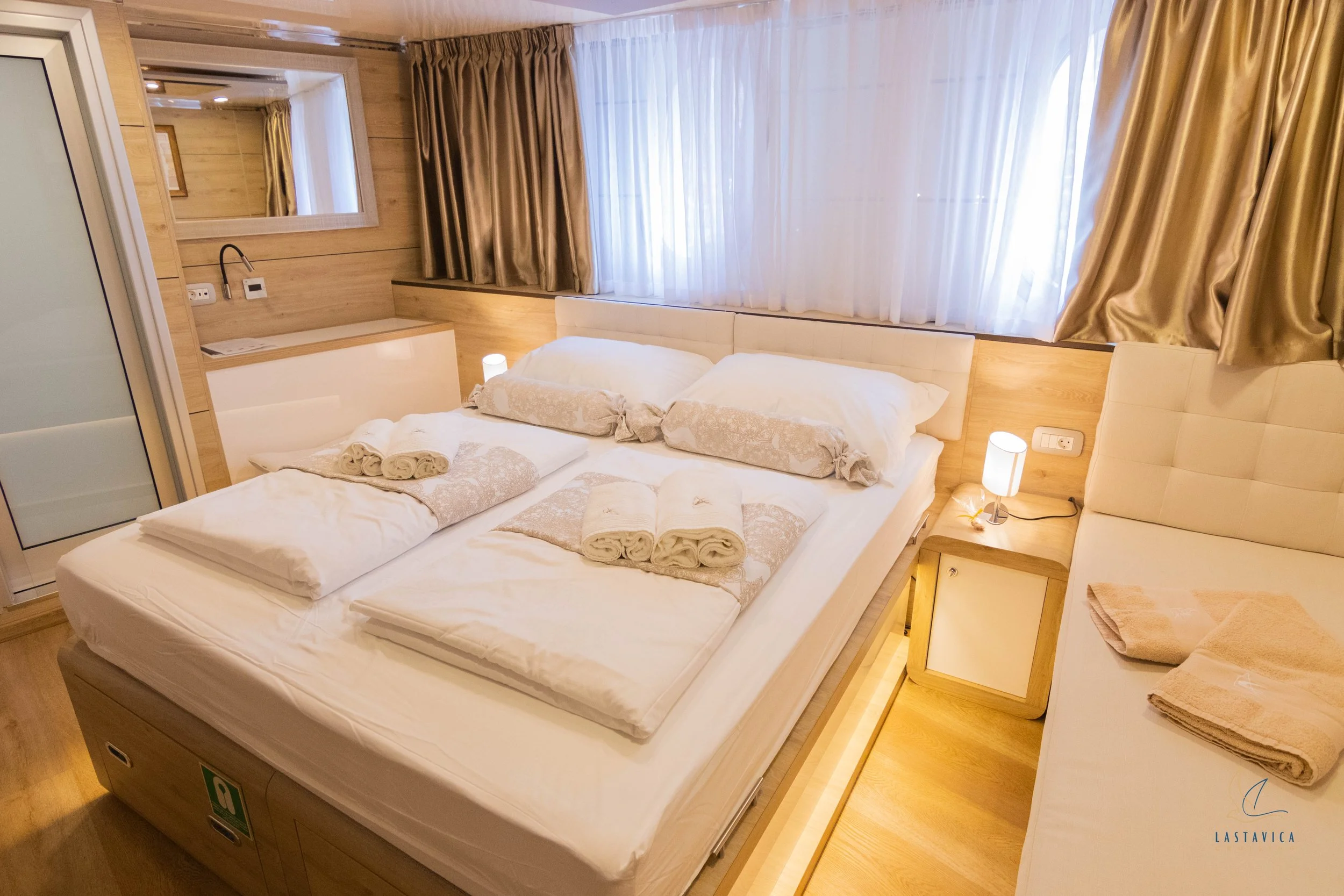A cozy hotel room with a bed, pillows, and rolled towels, featuring wooden accents and large windows with curtains.