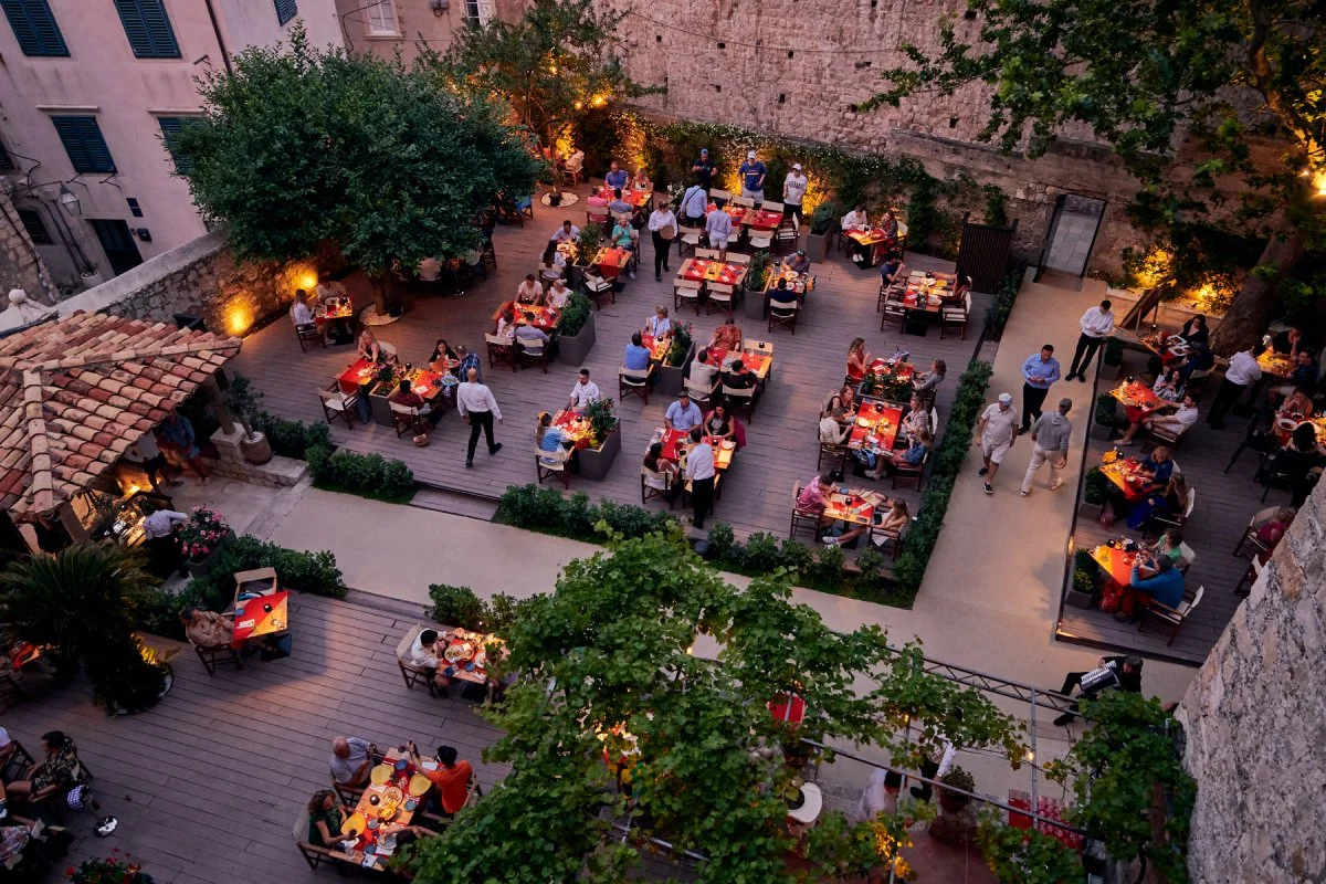 An aerial view of an outdoor restaurant patio at dusk, filled with tables of people dining, surrounded by trees and warm lighting. The restaurant is called Rudjer and located in Dubrovnik, Croatia