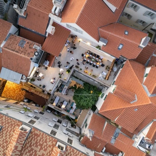 A rooftop dining and social gathering area with tables, chairs, umbrellas, and a large tree in the center, surrounded by red-tiled roofs of nearby buildings in an urban setting.