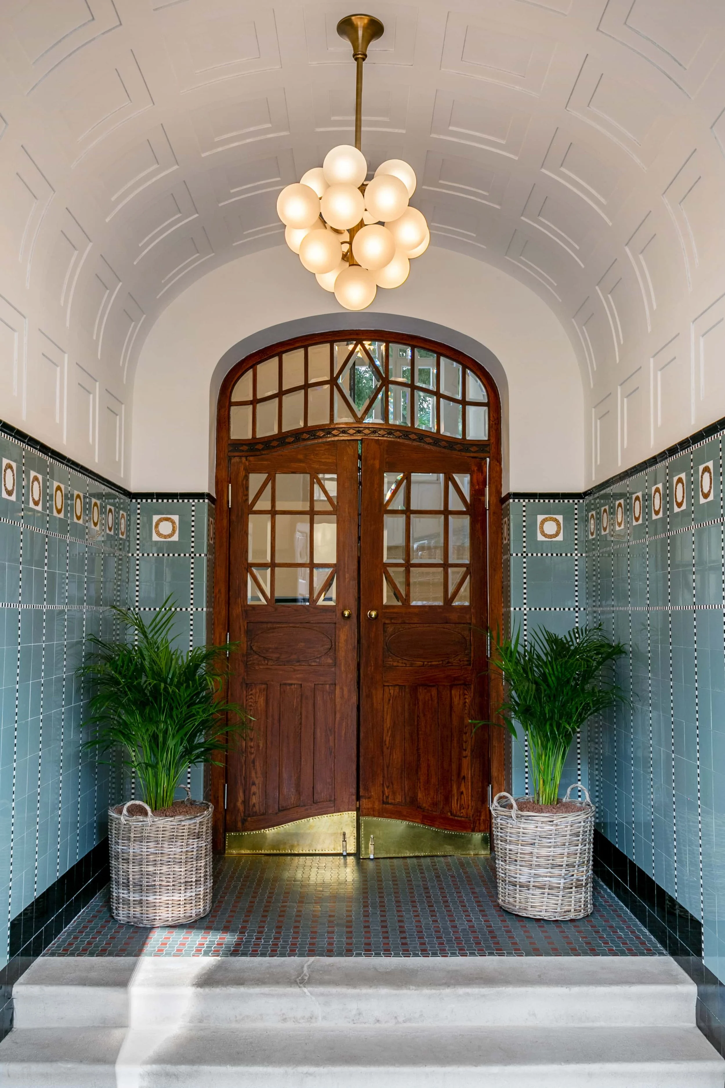 Decorative entryway with brass chandelier, wooden double doors with geometric grid pattern, green tile walls with circular accents, potted green plants in wicker baskets, and tiled floor with small geometric pattern, steps leading up to the door.