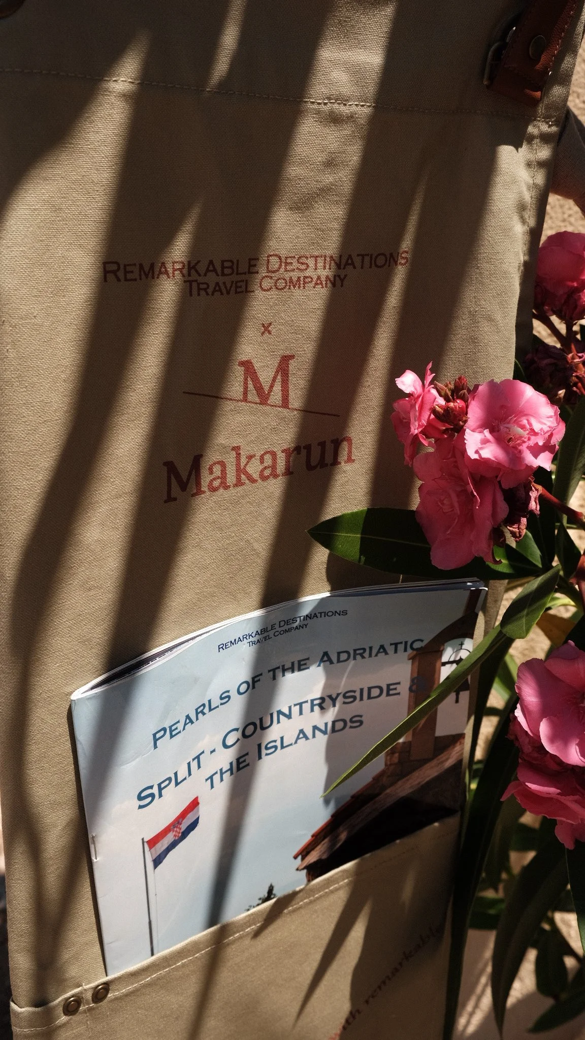 A travel brochure titled "Pearls of the Adriatic" is placed in a pocket of a beige bag, with pink flowers and a pink flower flower in front of it. The bag features the words "Remarkable Destinations Travel Company" and the letter "M".