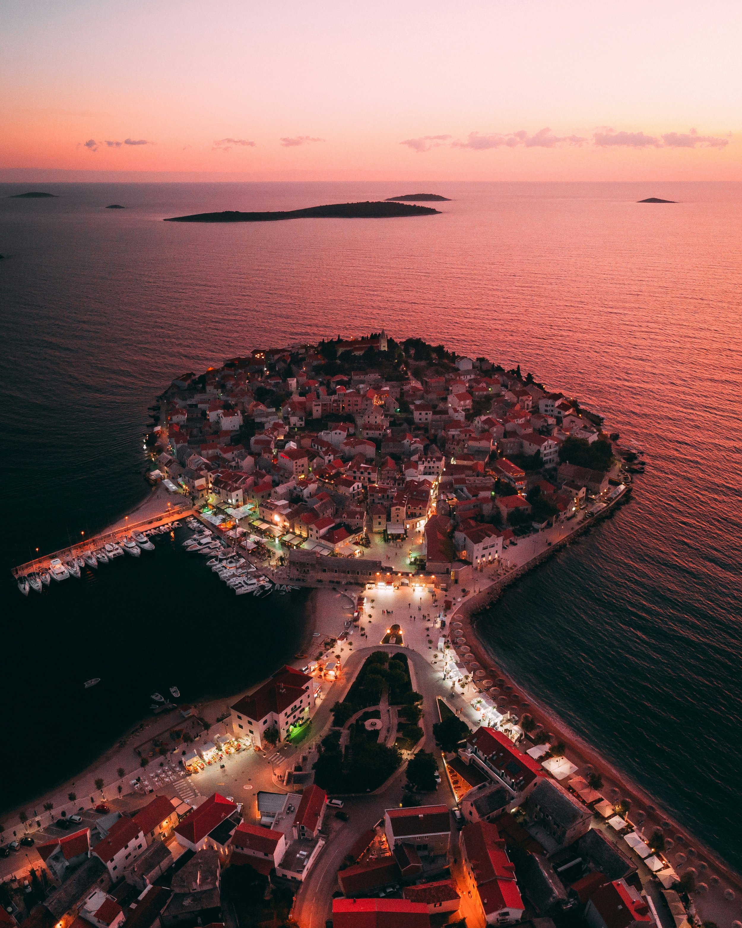 Aerial view of a coastal town during sunset, with a harbor filled with boats, narrow streets, and houses with red-tiled roofs, overlooking the ocean and small islands in the distance.