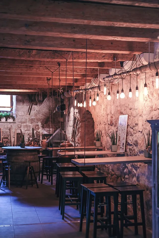 Empty rustic cafe with wooden tables and barstools, exposed brick walls, hanging bulbs, and potted plants on the ledge. Škar winery in Dubrovnik, Croatia