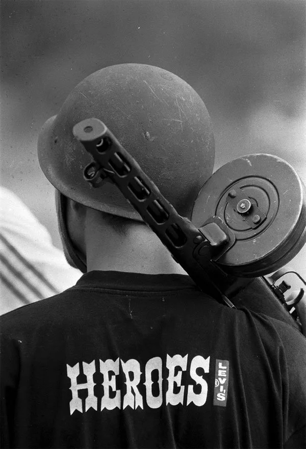 A person wearing a baseball cap and a t-shirt with the words 'Heroes' and 'Levi's' printed on the back, carrying a large camera or video equipment on their shoulder.