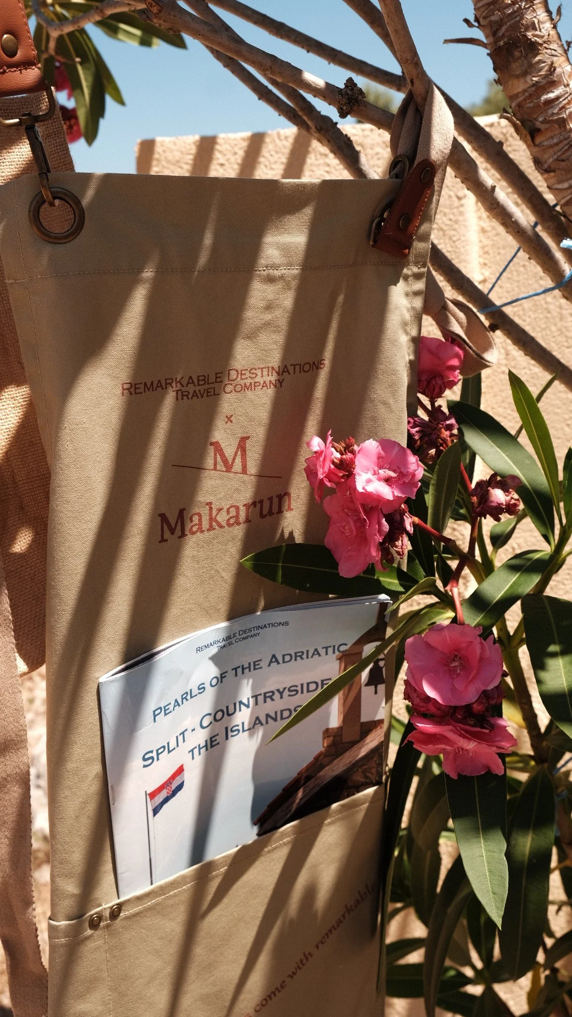 A travel brochure from Remarkable Destinations Travel Company attached to a beige fabric bag, with pink flowers and green leaves nearby. Shadow patterns cast by nearby bars are visible on the bag.