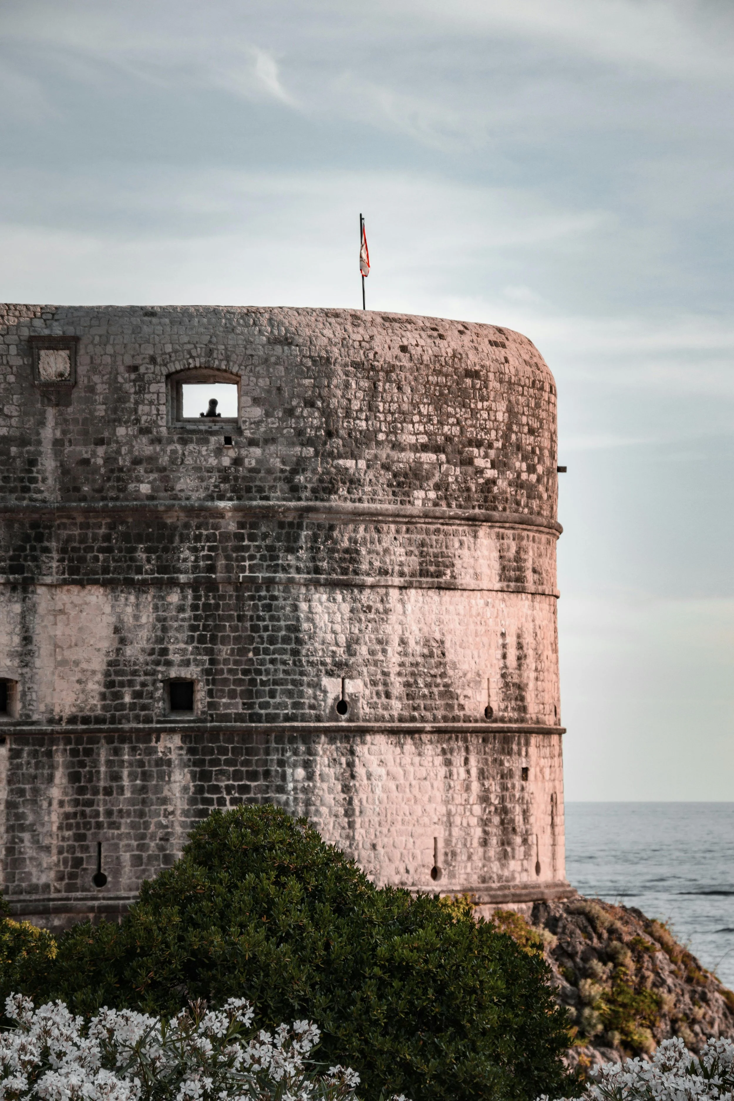 Old stone fortification or lighthouse on the coast with a red, white, and blue flag on top, overlooking the ocean. Green bushes and white flowers in the foreground, cloudy sky.