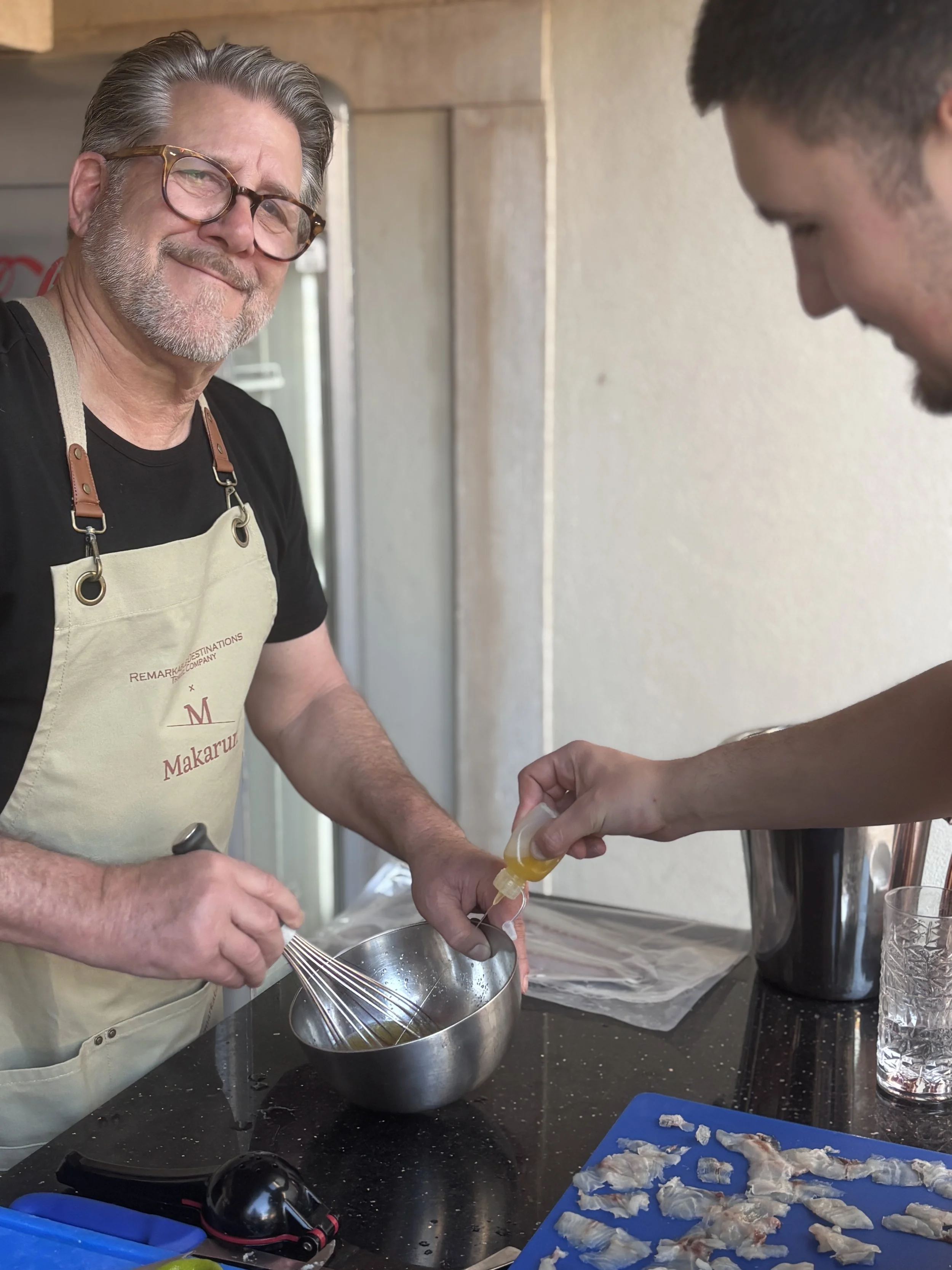 An older man with gray hair, wearing glasses and an apron, is holding a whisk and a mixing bowl while smiling at the camera. A younger person is pouring a liquid into the bowl. The scene appears to be a kitchen with various kitchen tools, a tray of r