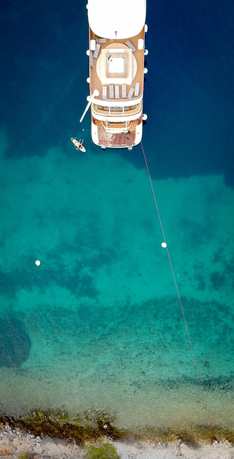 Overhead view of a luxury yacht called Lastavica anchored near a rocky Croatian shoreline with a small paddleboard and kayaker nearby in clear blue-green water.