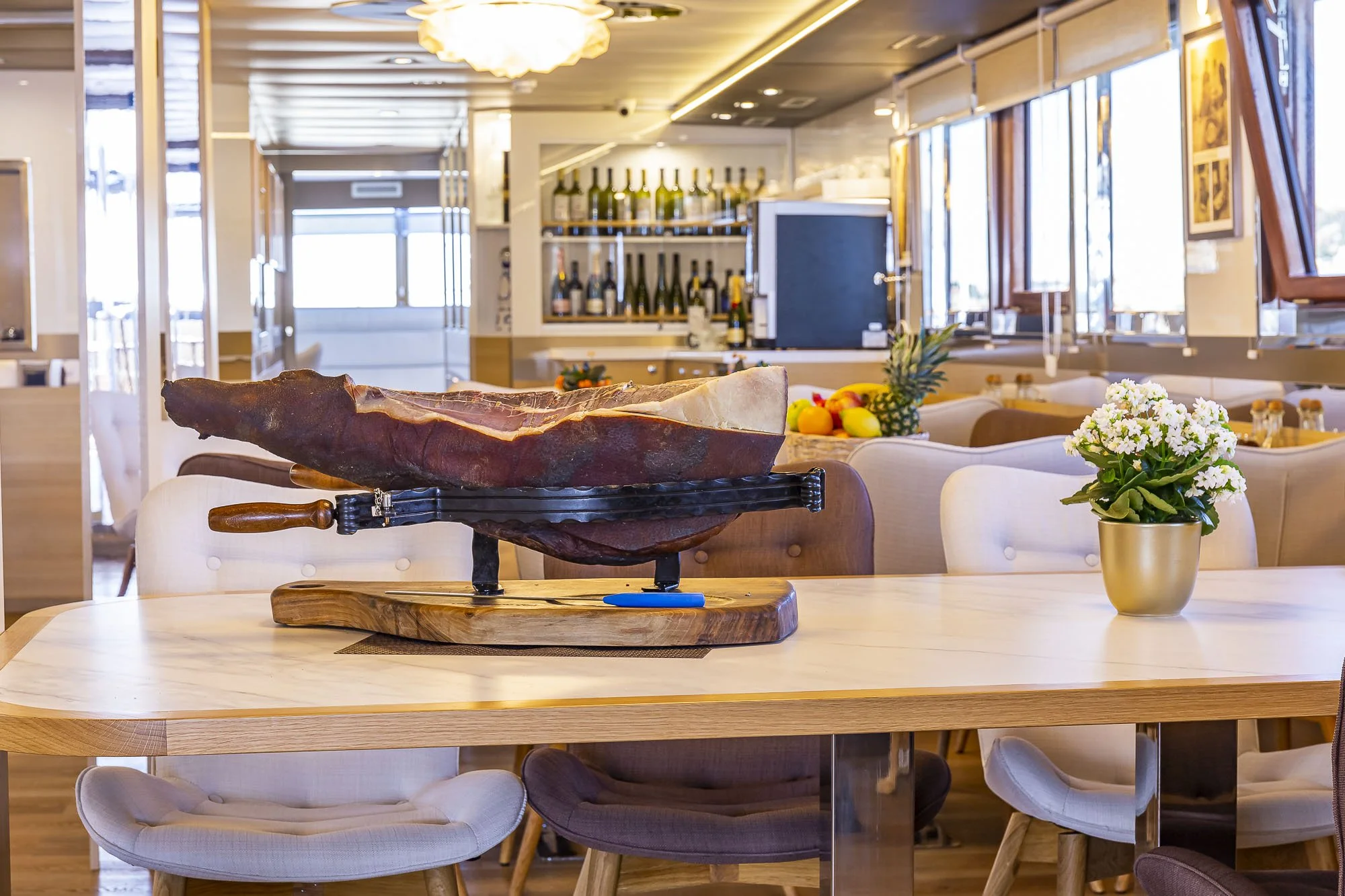 A restaurant with a wooden table setting, featuring a large piece of cured meat on a stand, a vase of white flowers, and a background of windows, shelves with wine bottles, and framed art.
