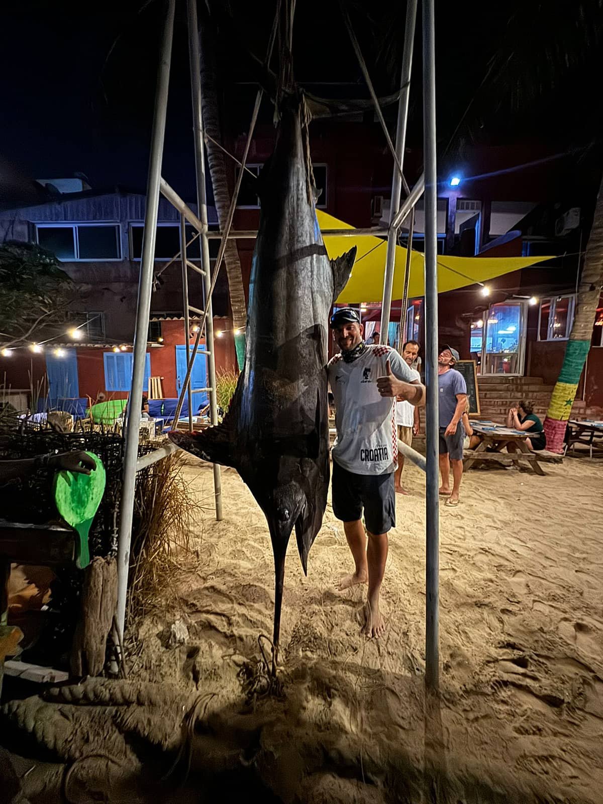 A man smiling and giving a thumbs-up next to a large, dark-colored marlin fish hanging vertically from a metal frame at a beachside restaurant during the evening. Several other people are seen in the background, and the setting includes sand, colorful umbrellas, string lights, and a building with large windows.