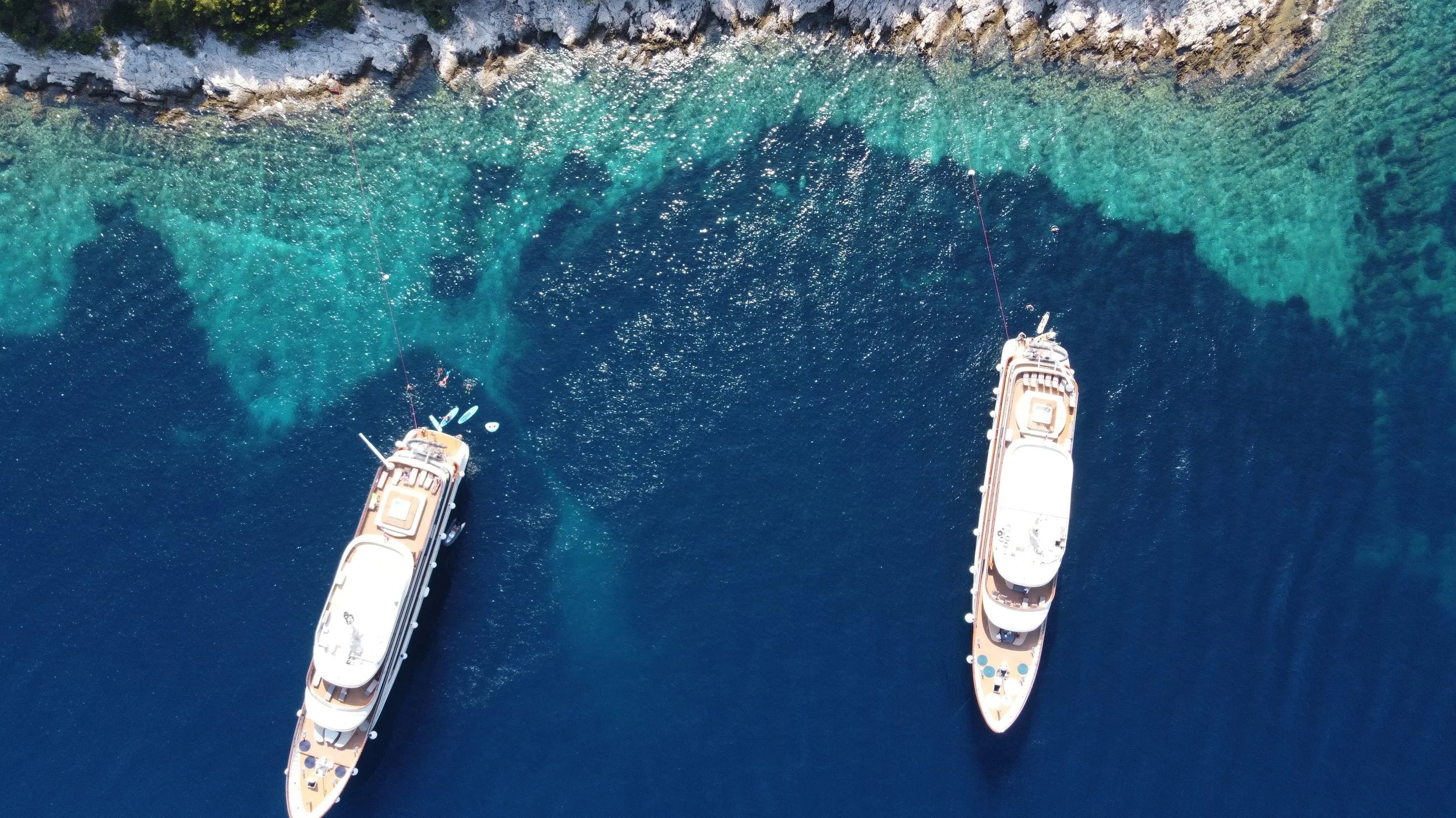 Aerial view of two yachts sailing in clear blue water near rocky shoreline.