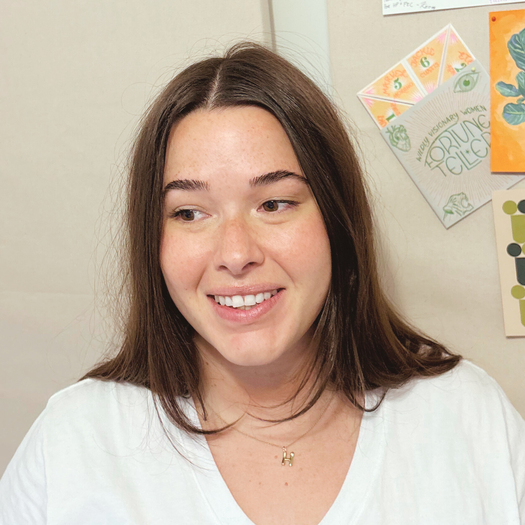 A young woman with long brown hair and freckles, smiling and wearing a white shirt and a gold necklace with the letter 'H'.