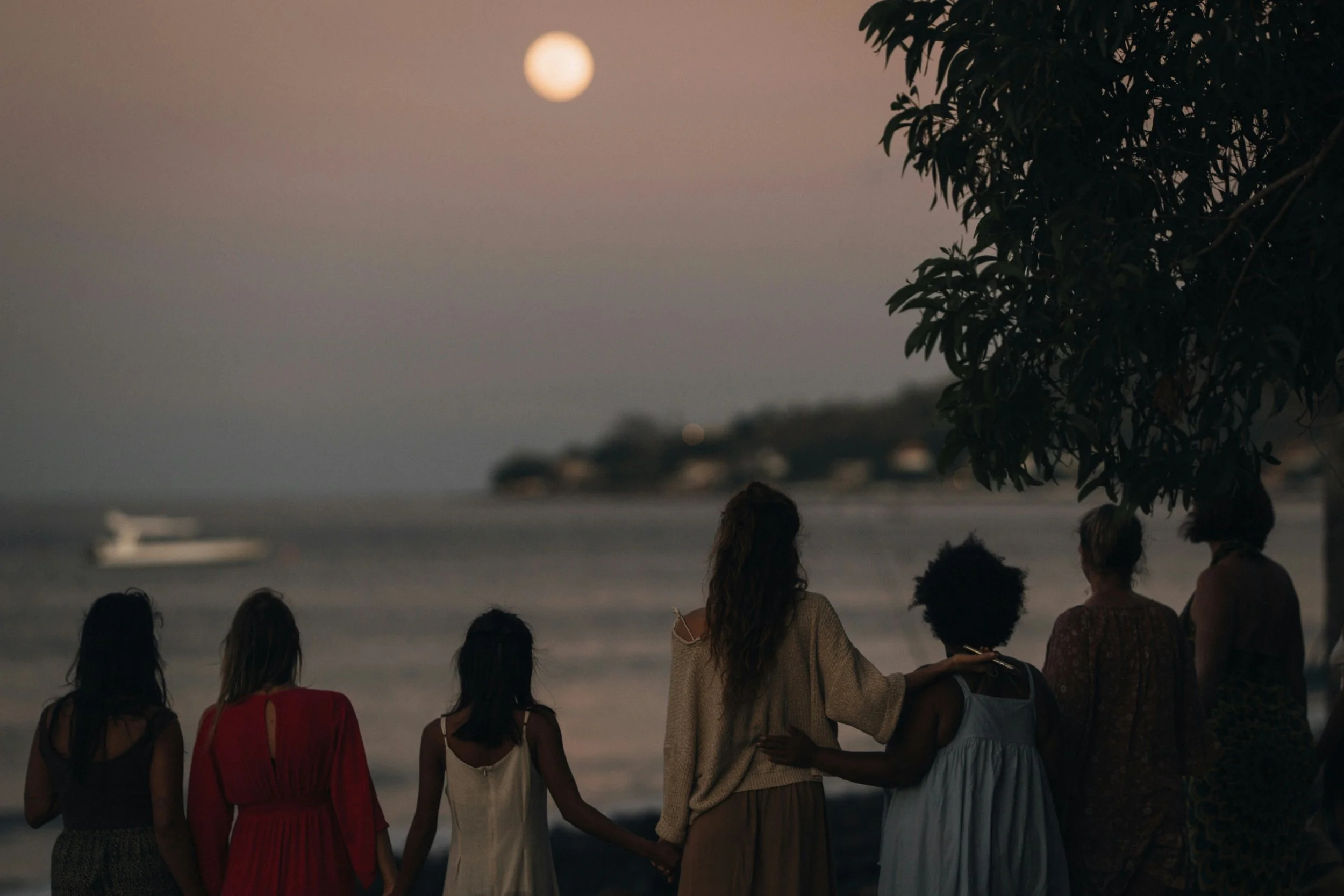 A group of people holding hands on a beach during sunset with the moon in the sky.
