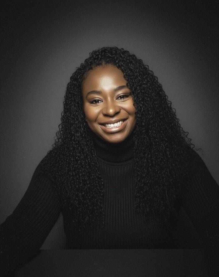 A woman with long curly black hair, smiling, wearing a black turtleneck, against a dark gray background.