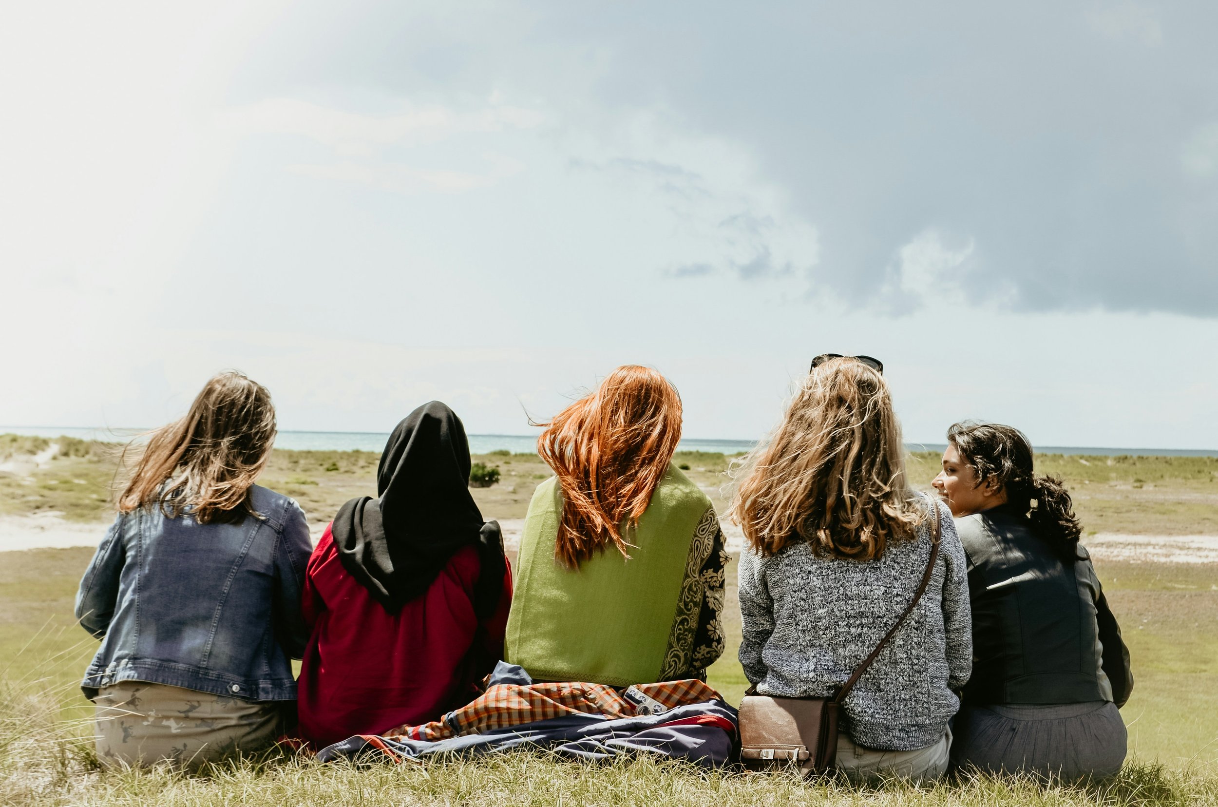 Five women sitting outdoors on the grass, facing away from the camera, overlooking a landscape with grass and water under a cloudy sky.