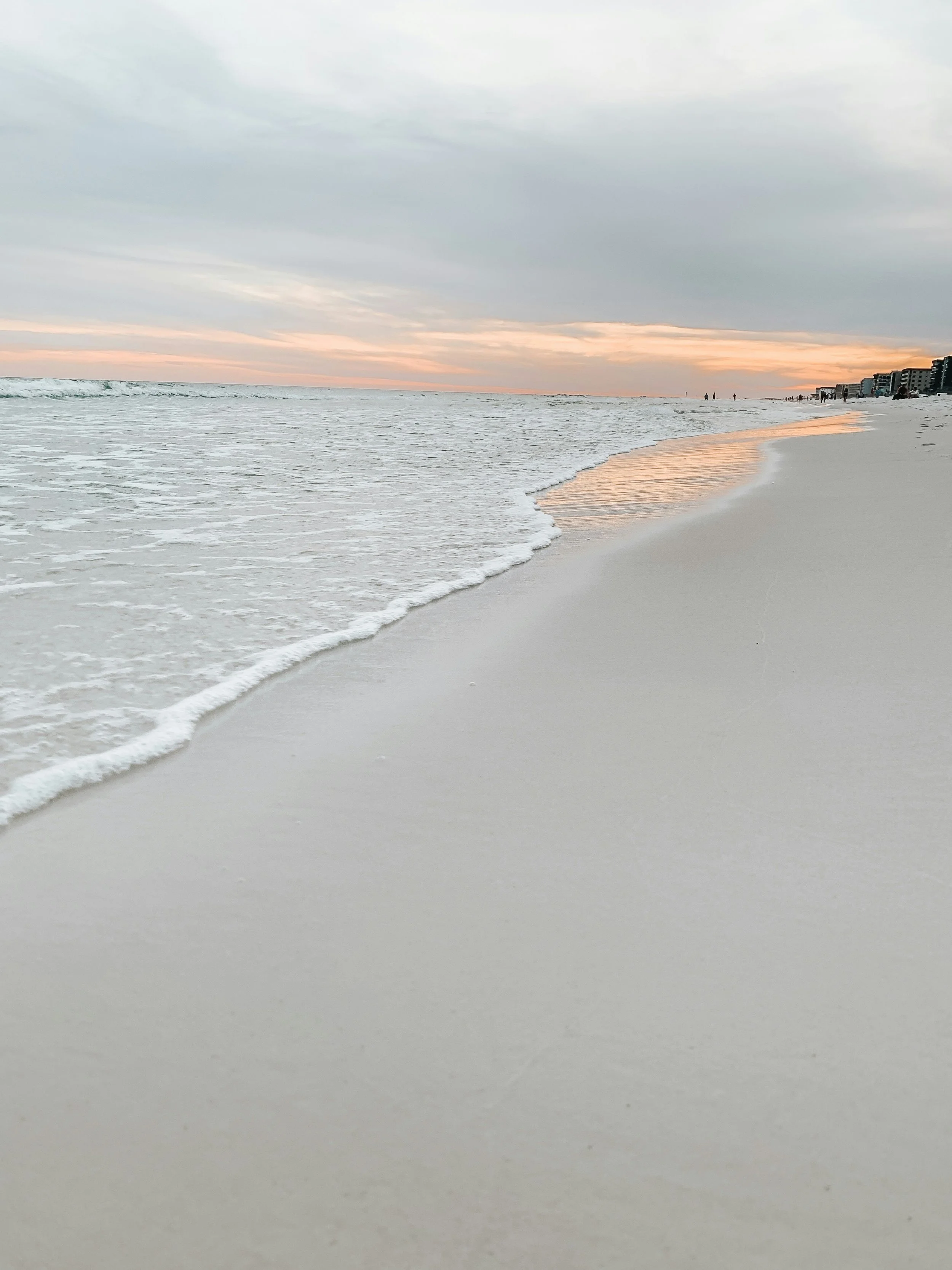 A sandy beach during sunset with gentle waves touching the shore and a row of buildings in the distance on the right.