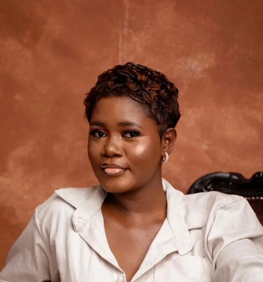 Portrait of a young woman with short, curly hair and earrings, sitting in front of a brown background.