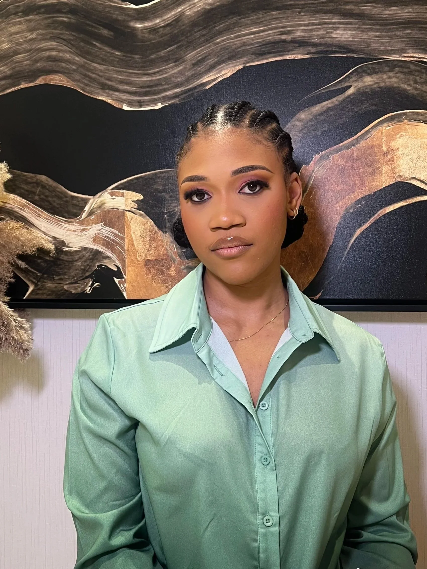 A woman with braided hair, makeup, and a light green button-up shirt standing in front of an abstract black and brown wall art.